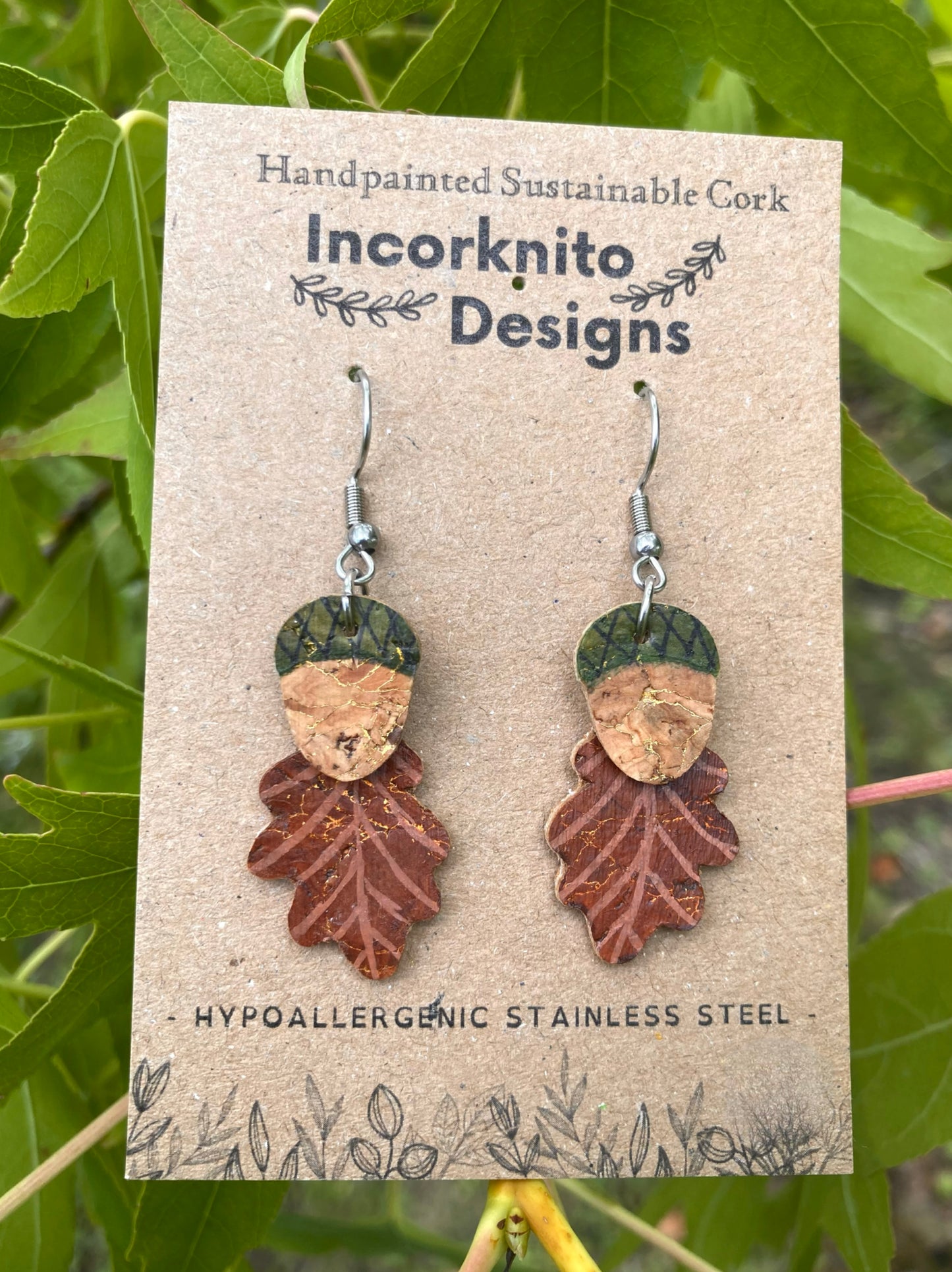 Pair of leaf-shaped earrings on a Incorknito Designs card with green leaves in the background