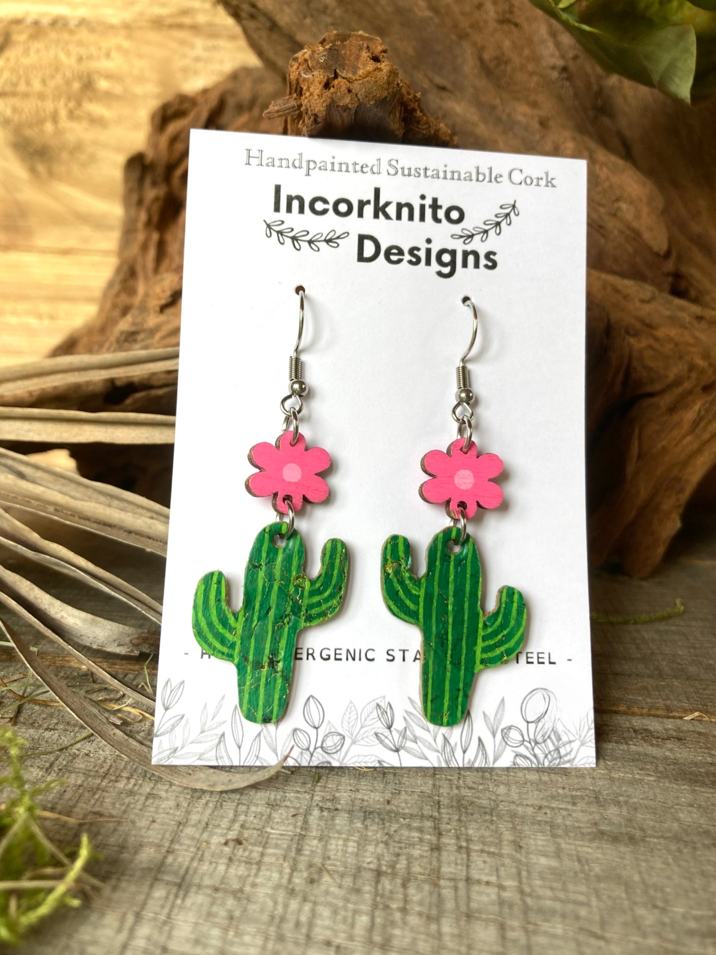 Cactus-shaped earrings with pink flowers on a 'Incorknito Designs' card, set against a natural wood background.