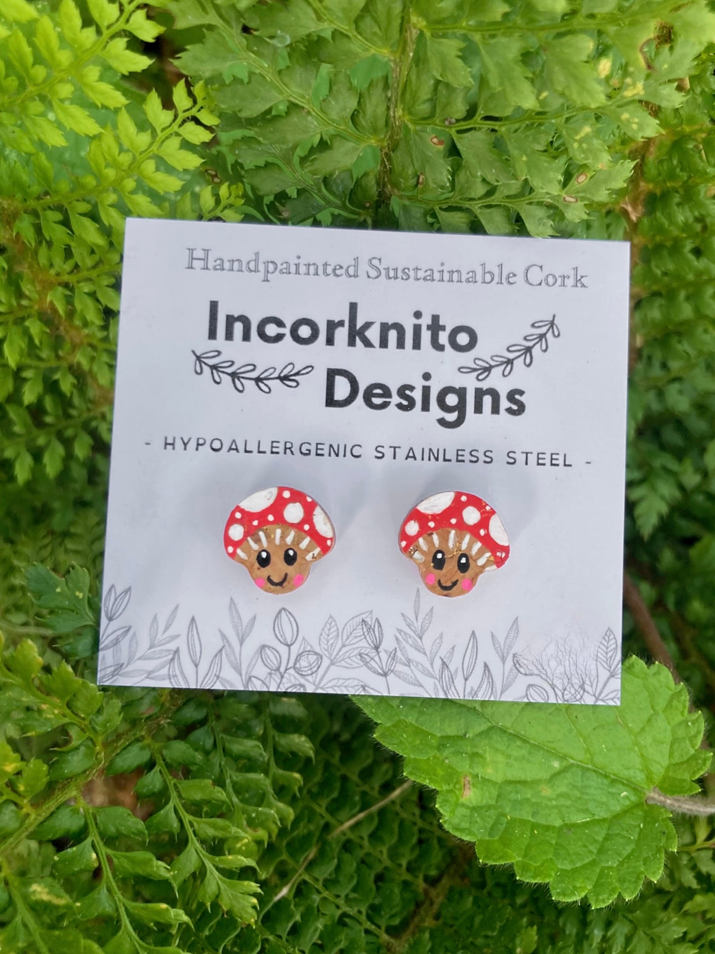 Toadstool-shaped earrings on a card with 'Incorknito Designs' branding, set against a green leafy background.