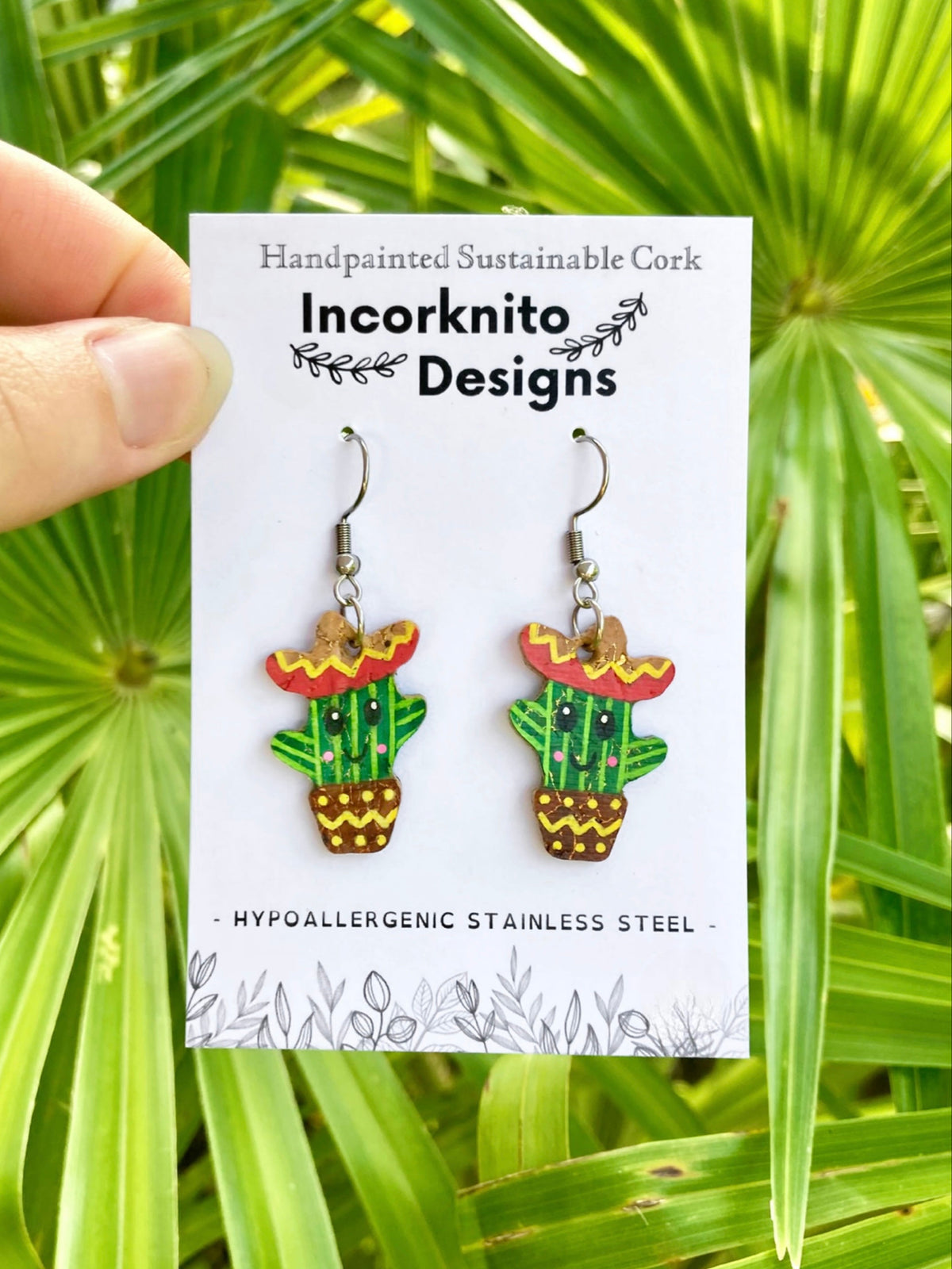 Happy cacti cork earrings on Incorknito Designs card, held against tropical palm leaves — hook fastening