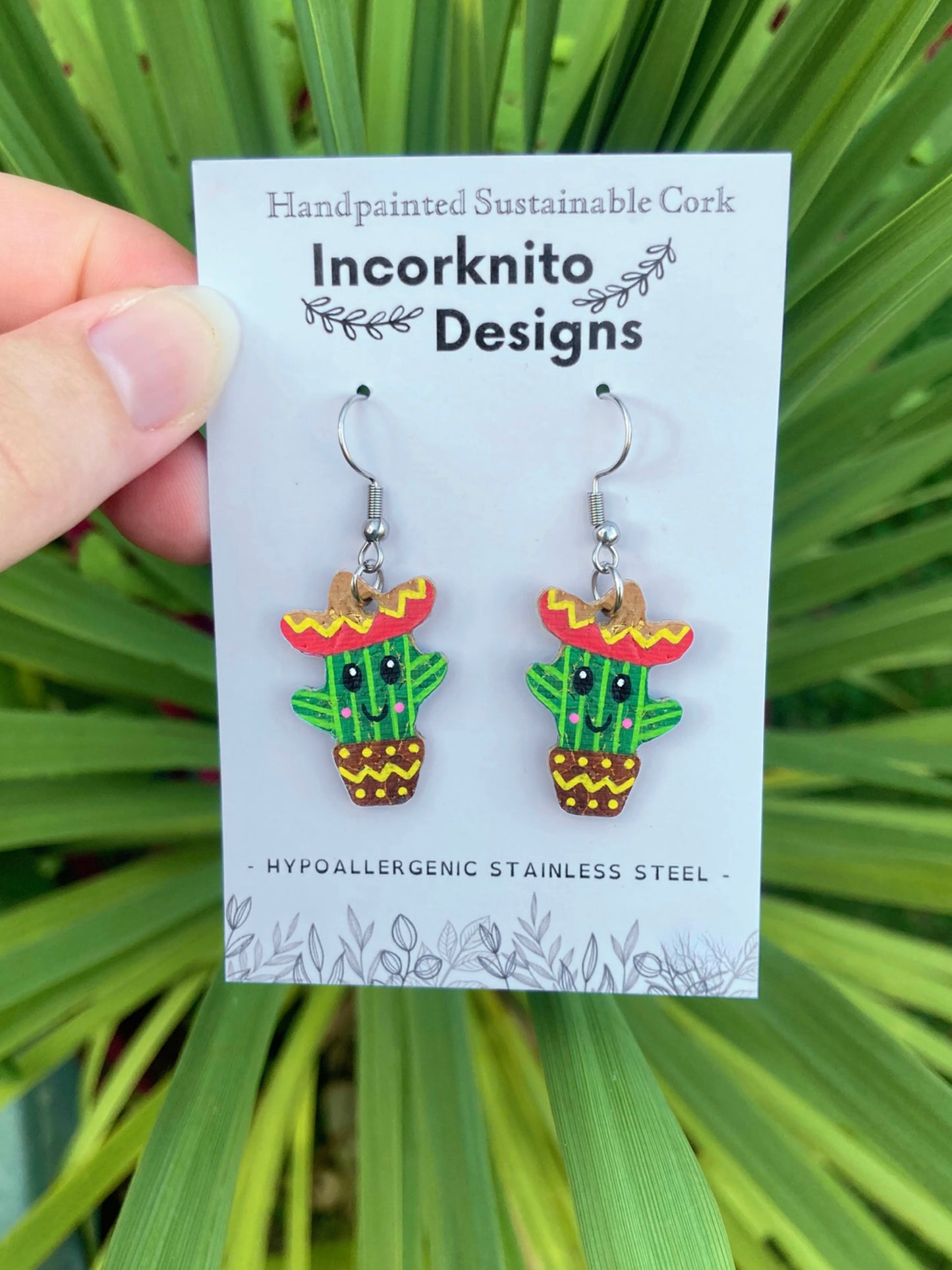 Cactus-shaped earrings on a card with 'Incorknito Designs' branding against a green leafy background.