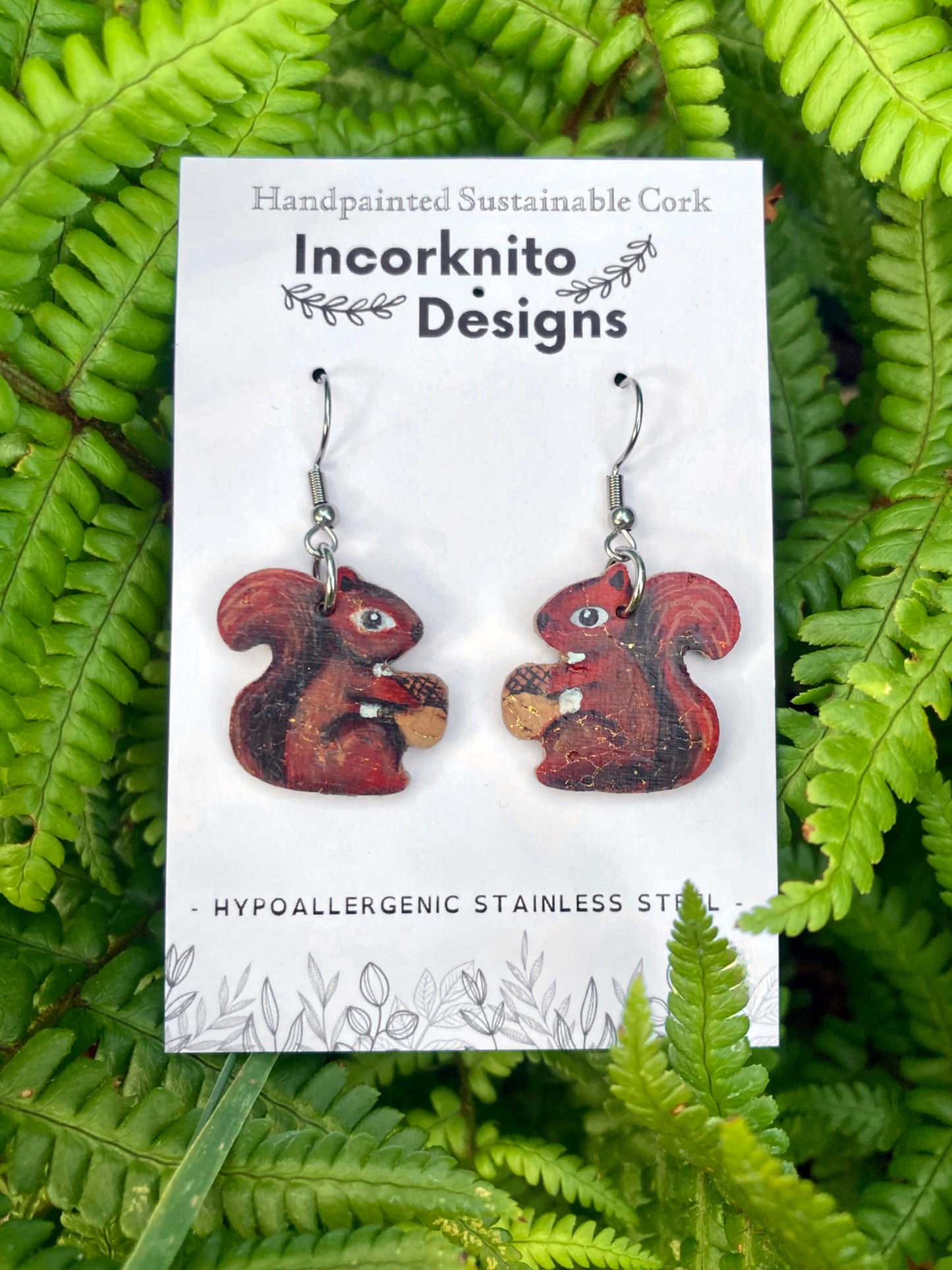 Squirrel-shaped earrings on a card with 'Incorknito Designs' branding, set against a green leafy background.