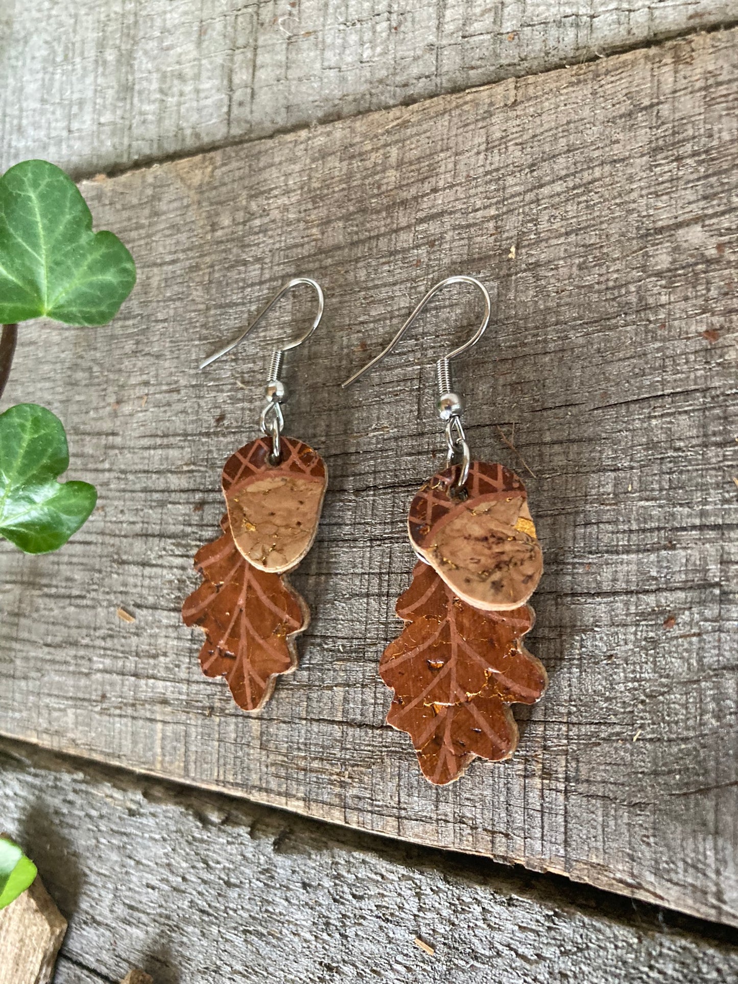 A pair of brown cork leather acorn and oak leaf dangle earrings with gold flecks, displayed against a wooden background.