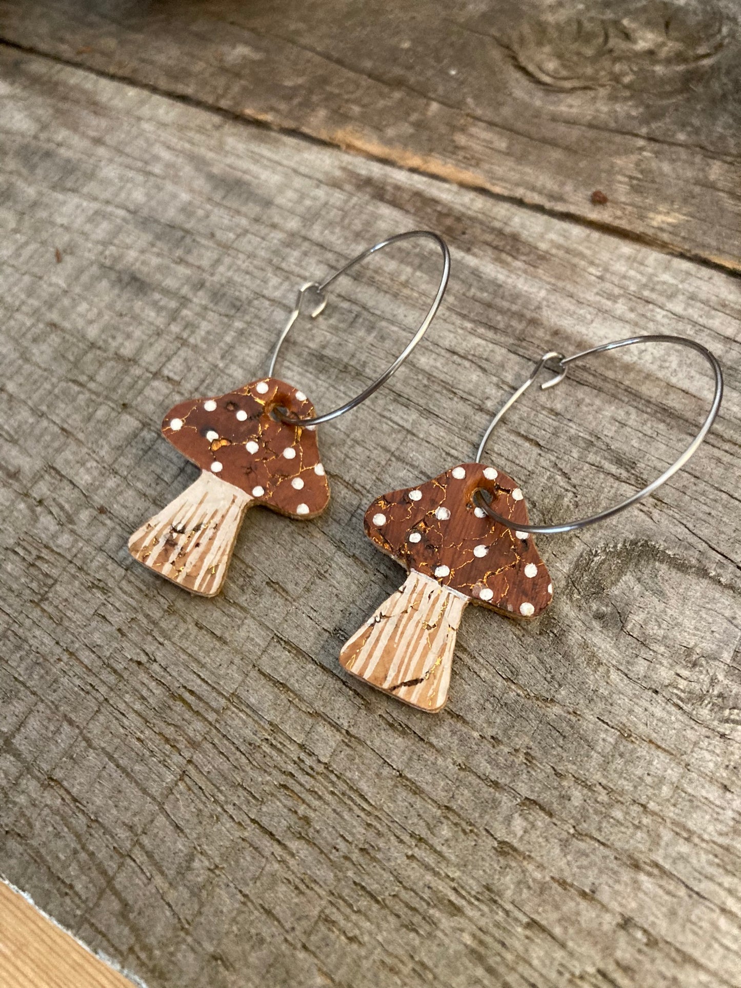 A pair of brown toadstool-shaped hoop earrings with gold flecks, made from sustainable cork, displayed against a wooden background.