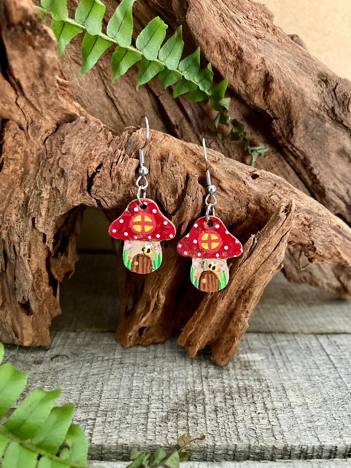 A pair of red fairy house toadstool earrings, featuring a cute tiny bee, a door, and a garden. Made from sustainable cork leather with gold flecks.