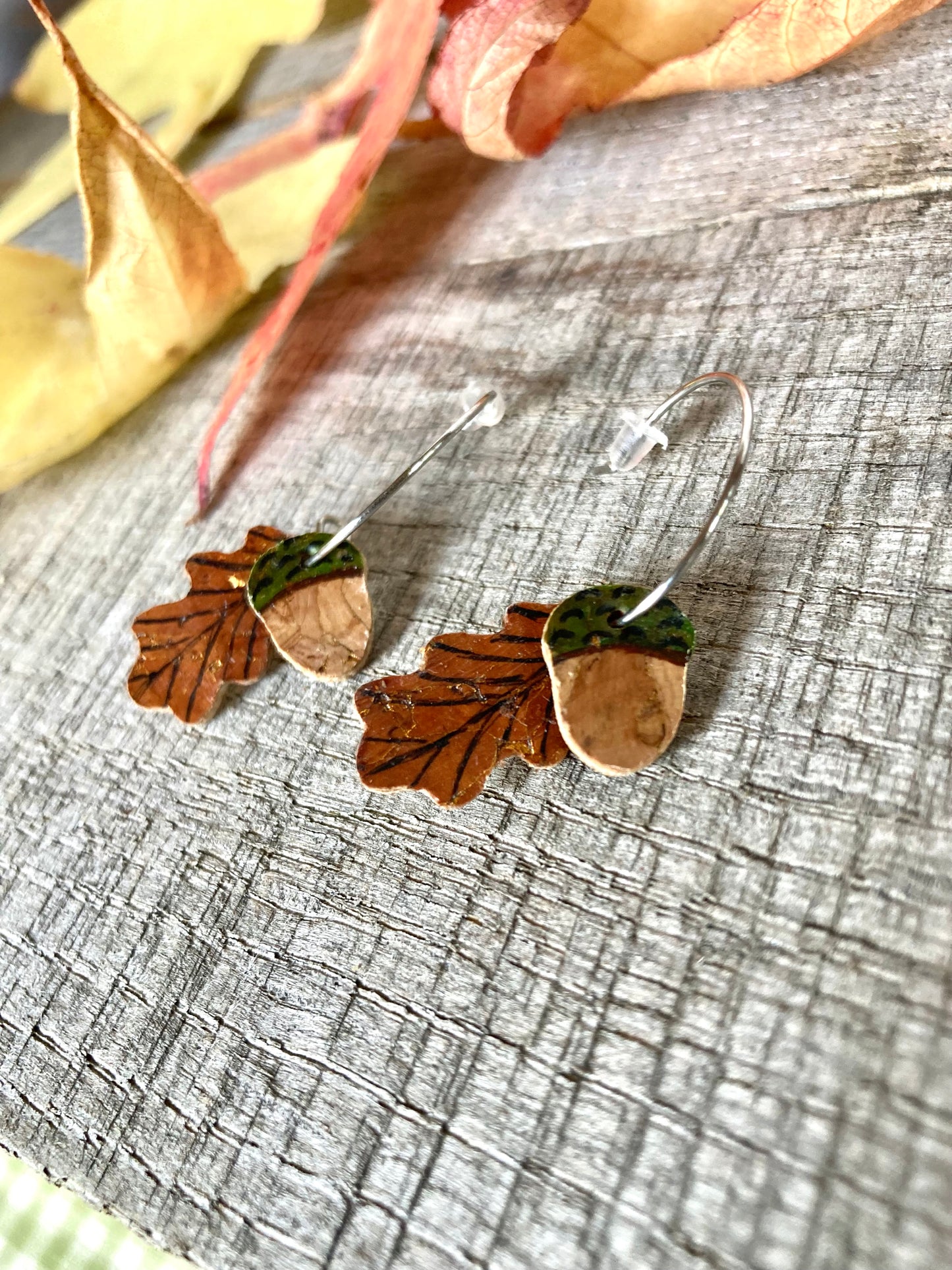 A pair of hand-painted cork leather mini hoop earrings with acorn and oak leaf designs, displayed on a wooden surface with autumn leaves scattered around.