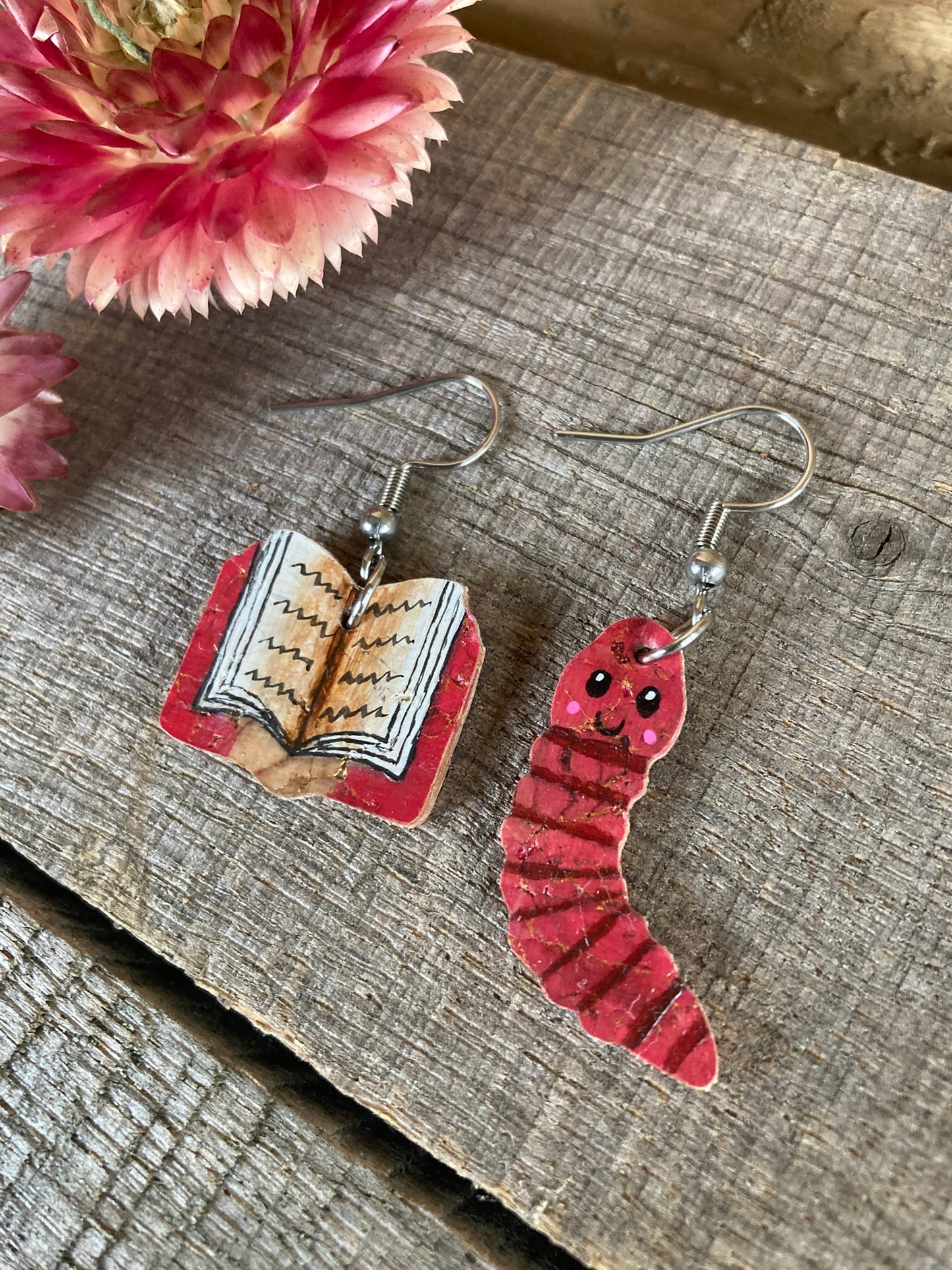 Two mismatched earrings - one book and one pink worm on a wooden surface with a pink flower in the background