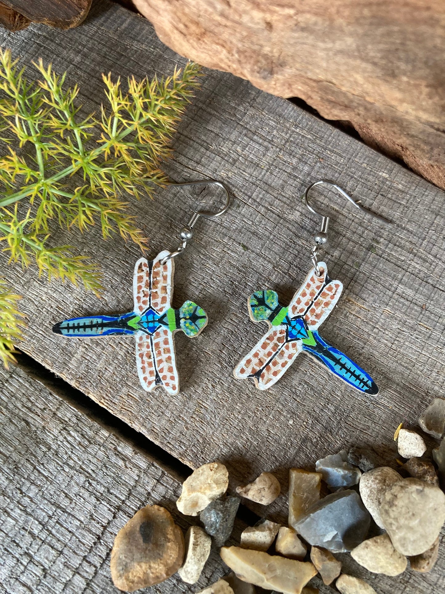 Dragonfly-shaped earrings on a wooden surface with greenery and stones.