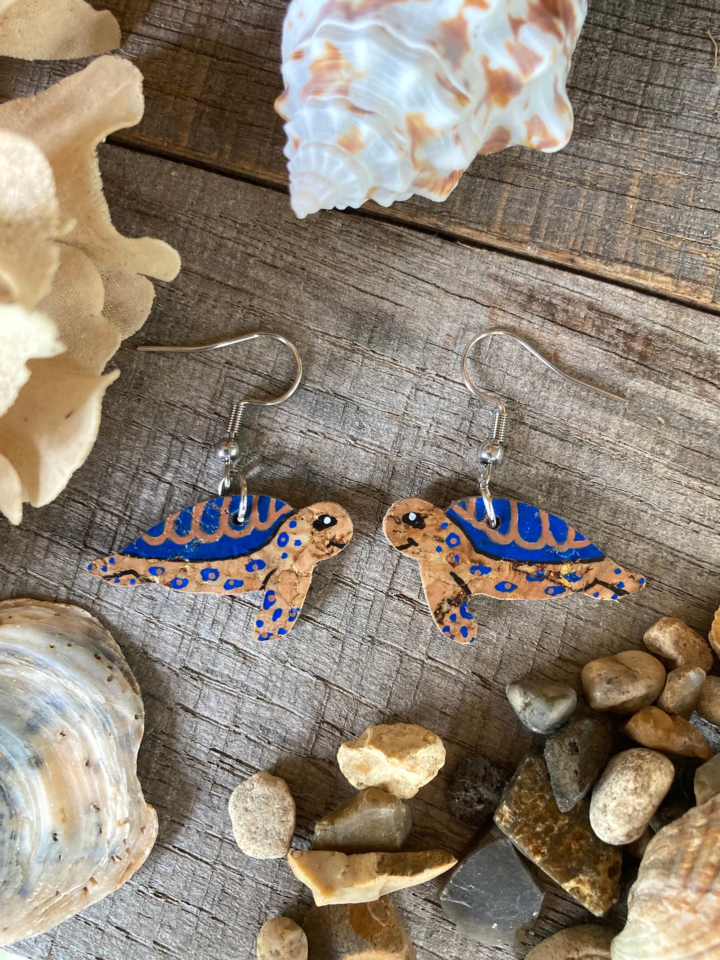 Turtle-shaped earrings with blue accents on a wooden surface with seashells and stones.
