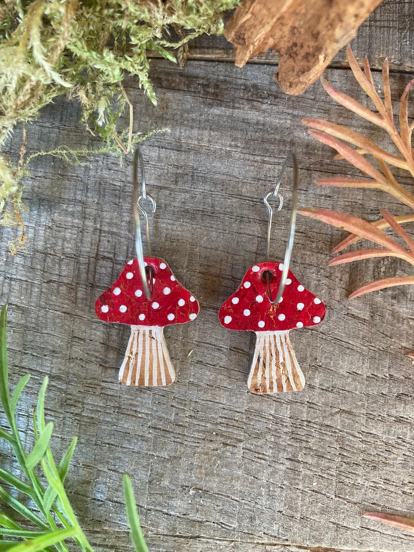 Red mushroom-shaped earrings with white polka dots on a wooden surface with greenery.