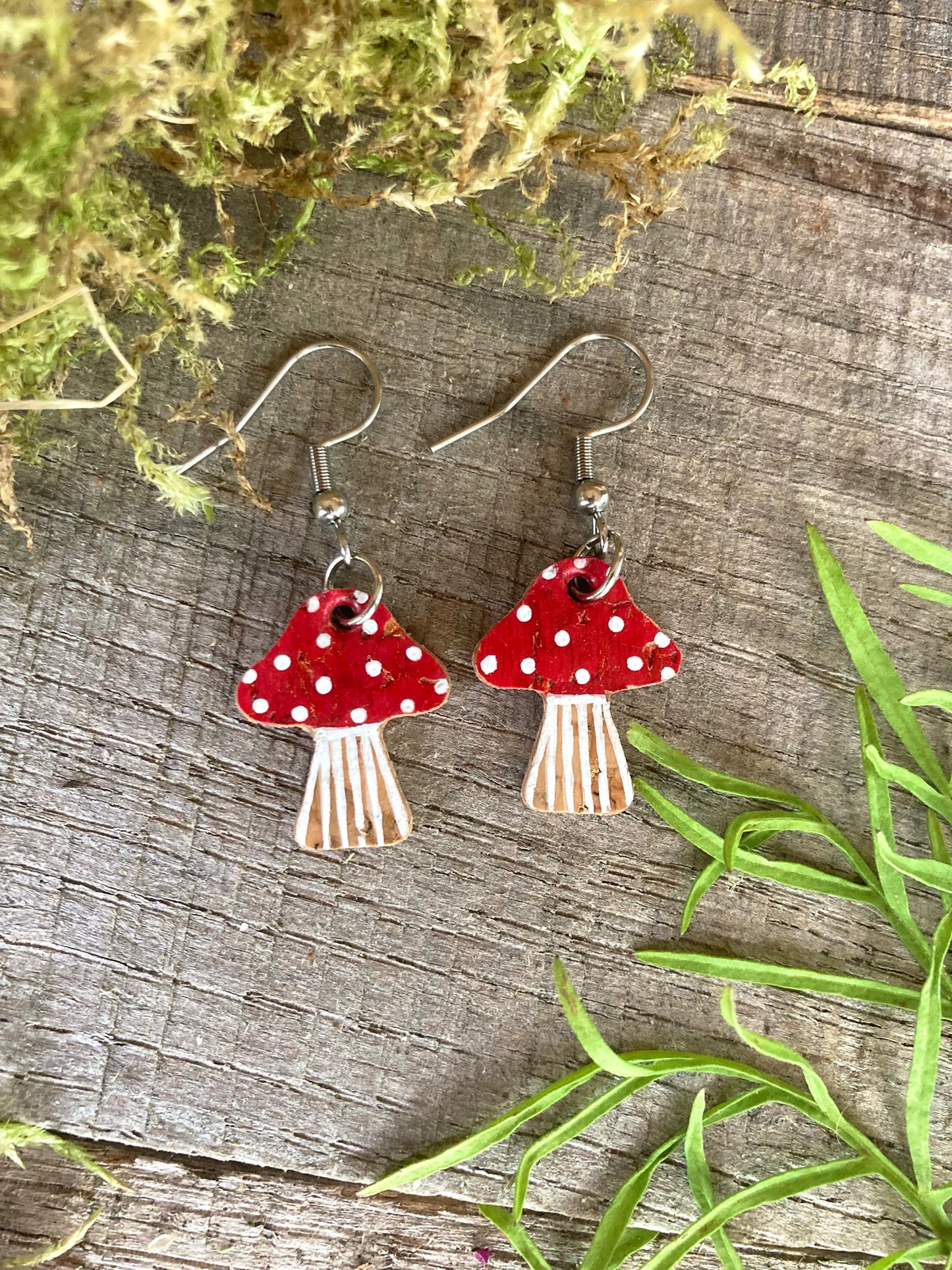 Red and white mushroom-shaped earrings on a wooden surface with greenery.