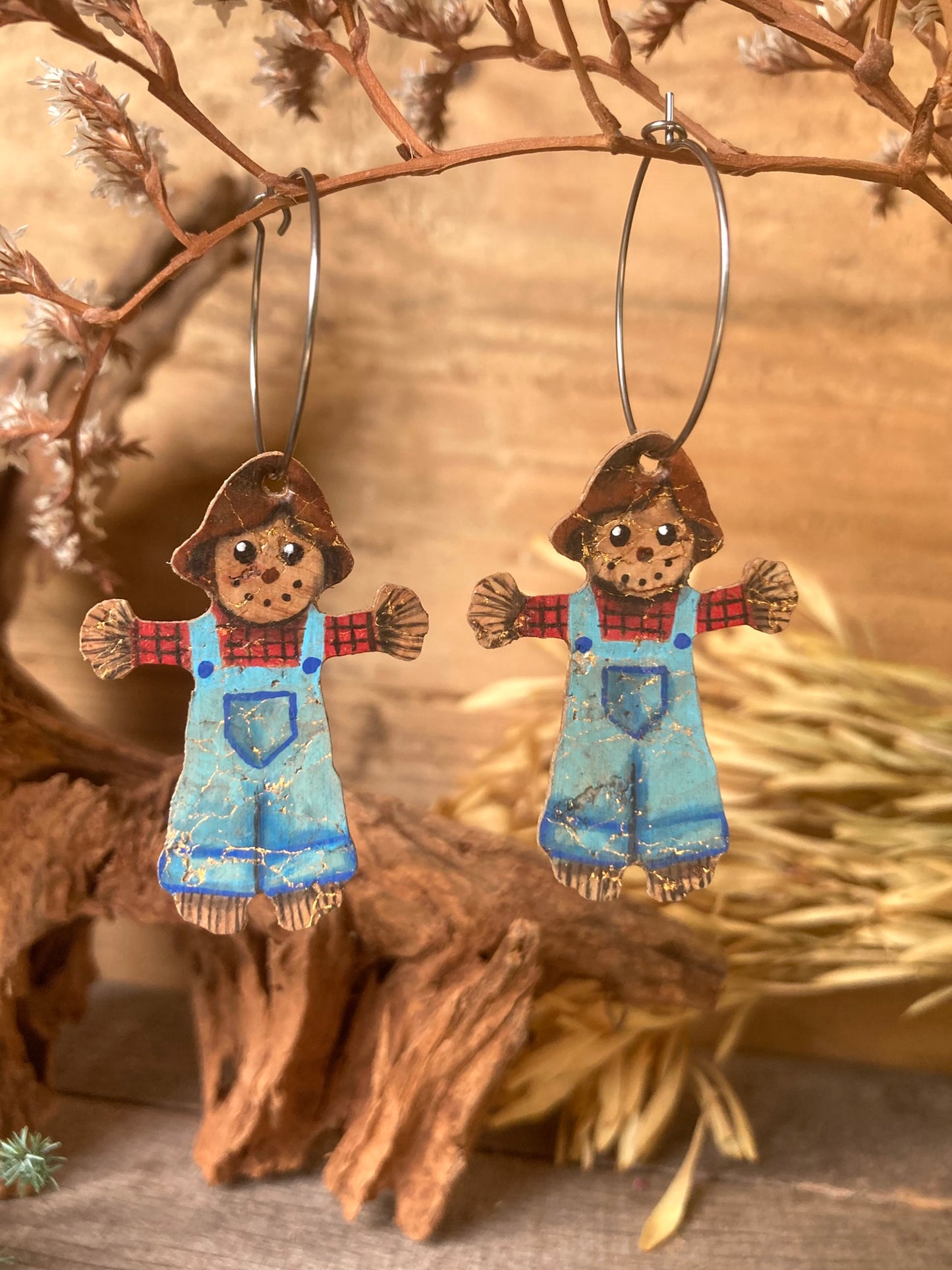 Scarecrow-themed earrings hanging on a branch with a natural background. Gold flecks in the cork earrings glisten in the sunlight
