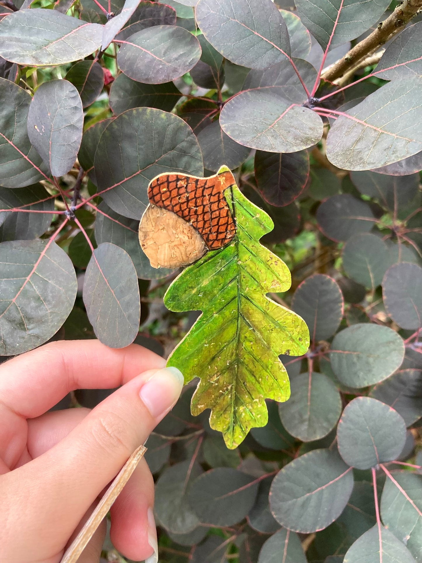 Hand holding a green leaf-shaped bookmark with an acorn against a background of green leaves.