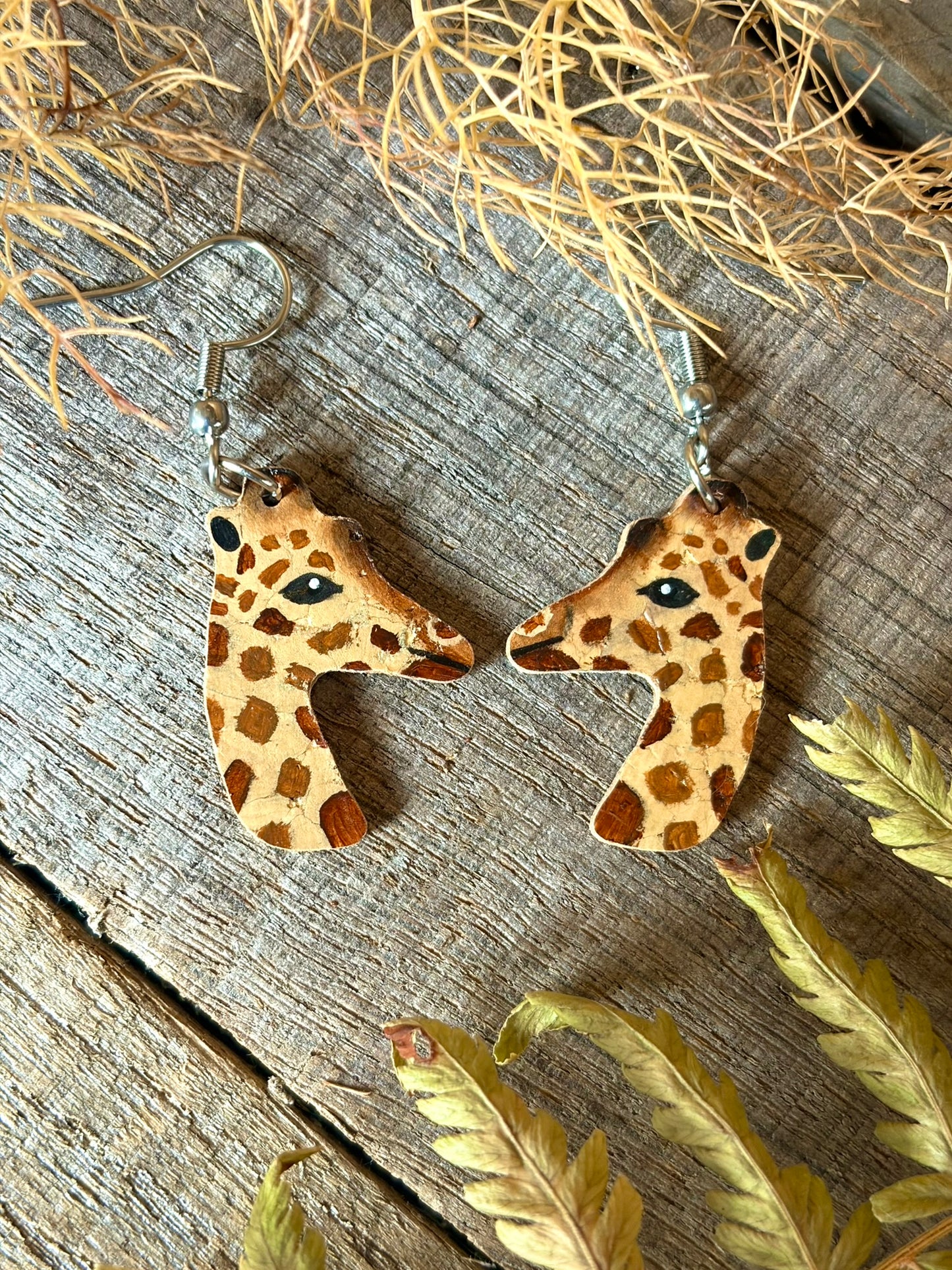 A pair of giraffe head dangle earrings with a natural wood and tan pattern, displayed on a rustic wooden surface with dried ferns.