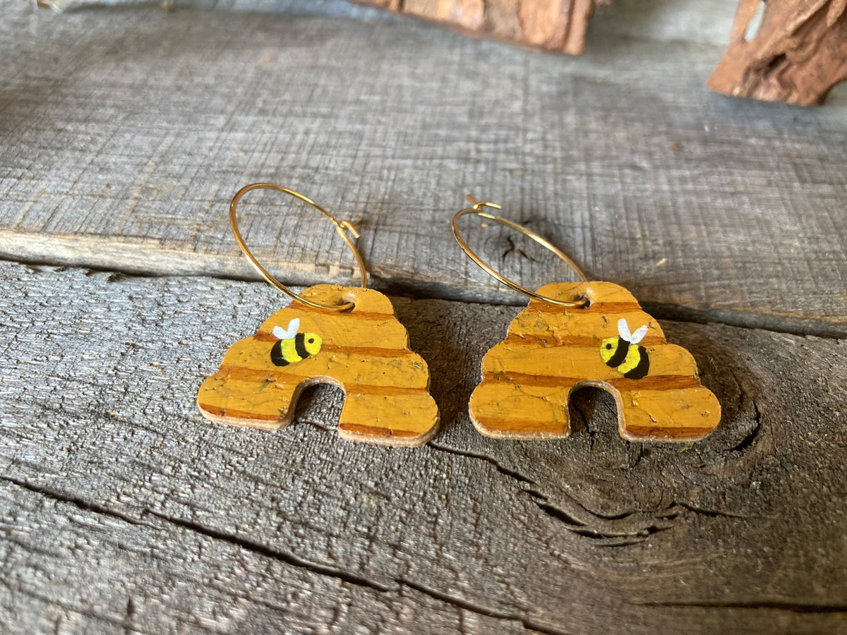 Handpainted cork beehive earrings on gold hoops, close up flat lay on weathered wood showing bumblebee and horizontal band detail
