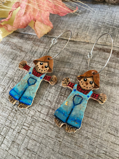 A pair of handcrafted scarecrow earrings with blue painted dungarees and gold-coloured details on a wooden background, displayed on a rustic textured surface with autumn leaves.