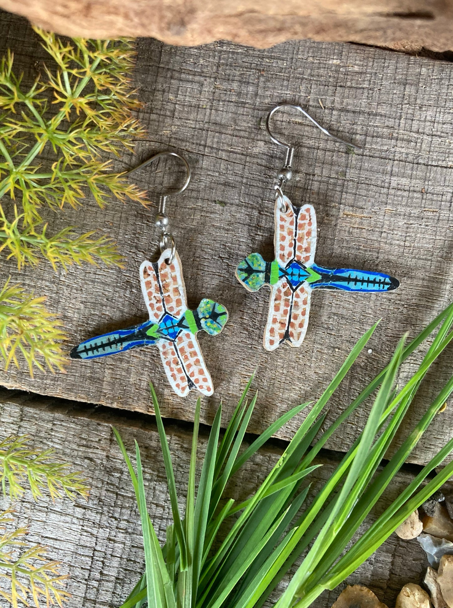 Dragonfly-shaped earrings on a wooden surface with greenery