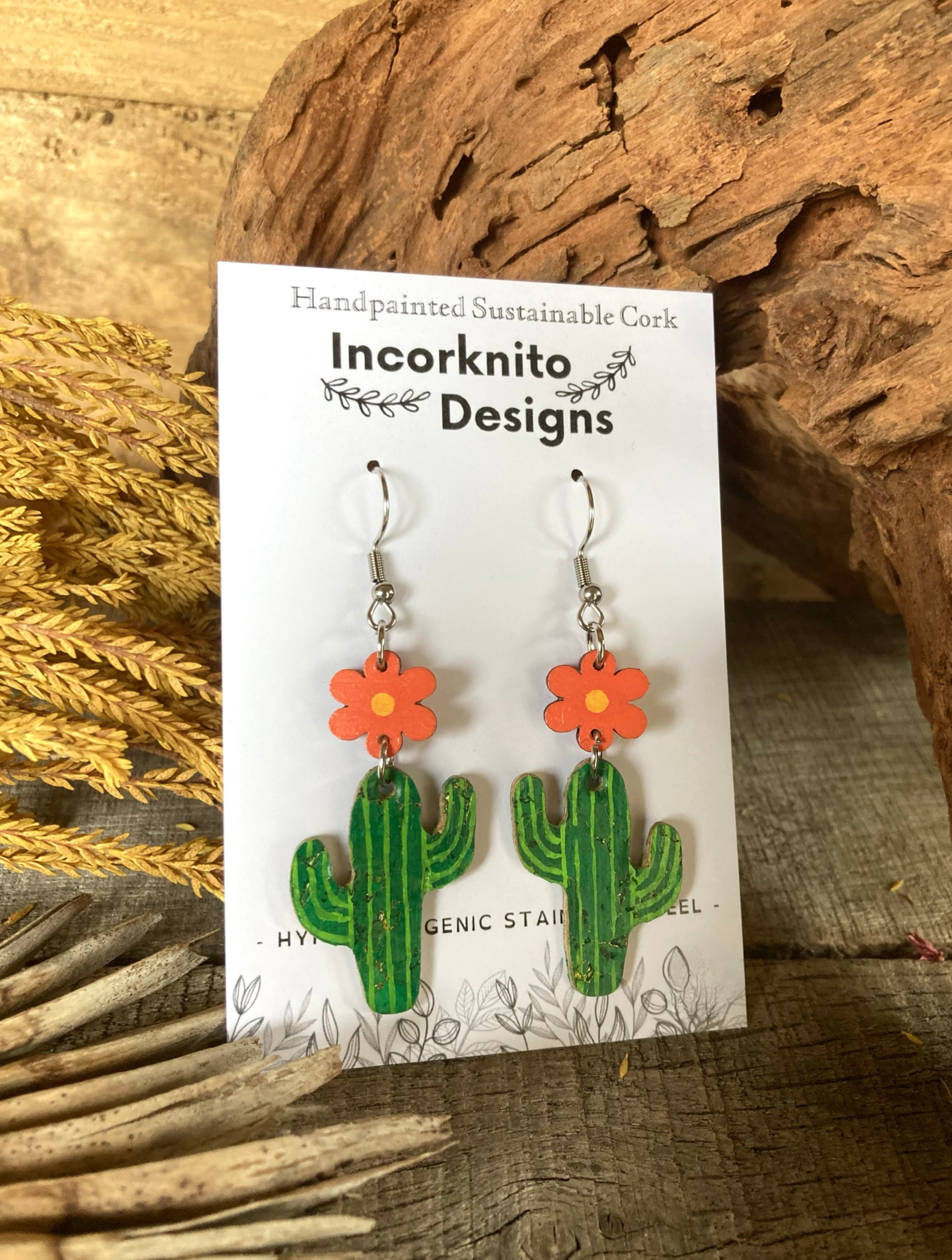 Cactus-shaped earrings with orange flowers on a 'Incorknito Designs' card, set against a natural wood background.