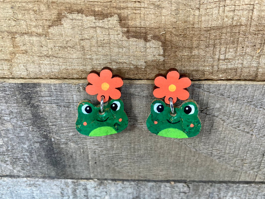 Happy faced frog earrings hanging from a orange flower stud on a wooden background. Made from gold flecked sustainable cork