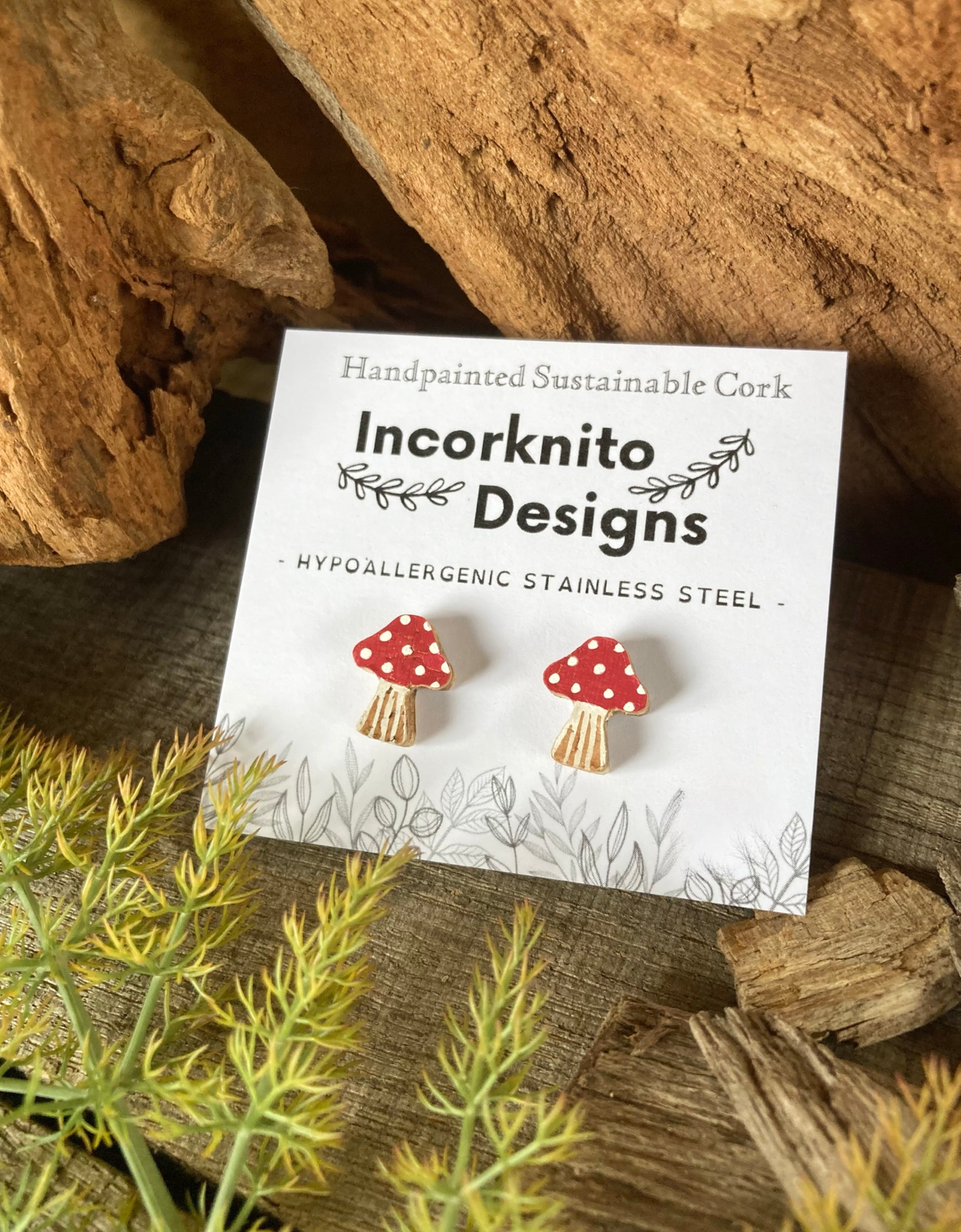 Red and white mushroom-shaped earrings on a card with 'Incorknito Designs' branding, placed on a natural wood and plant background.