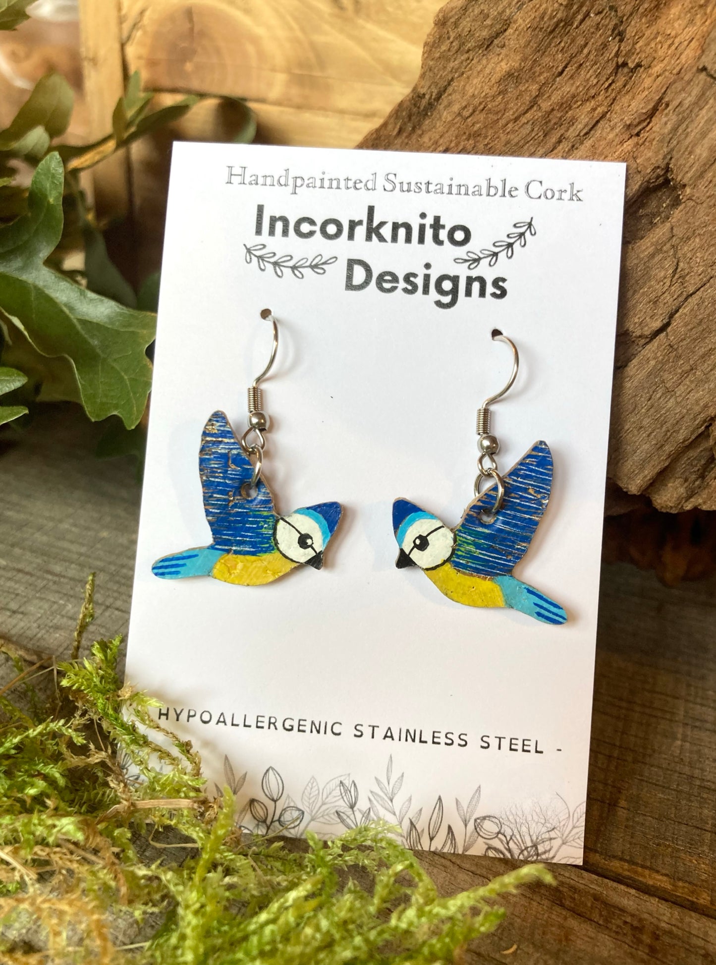 Bird-shaped earrings on a card with 'Incorknito Designs' branding, placed on a wooden surface with greenery.