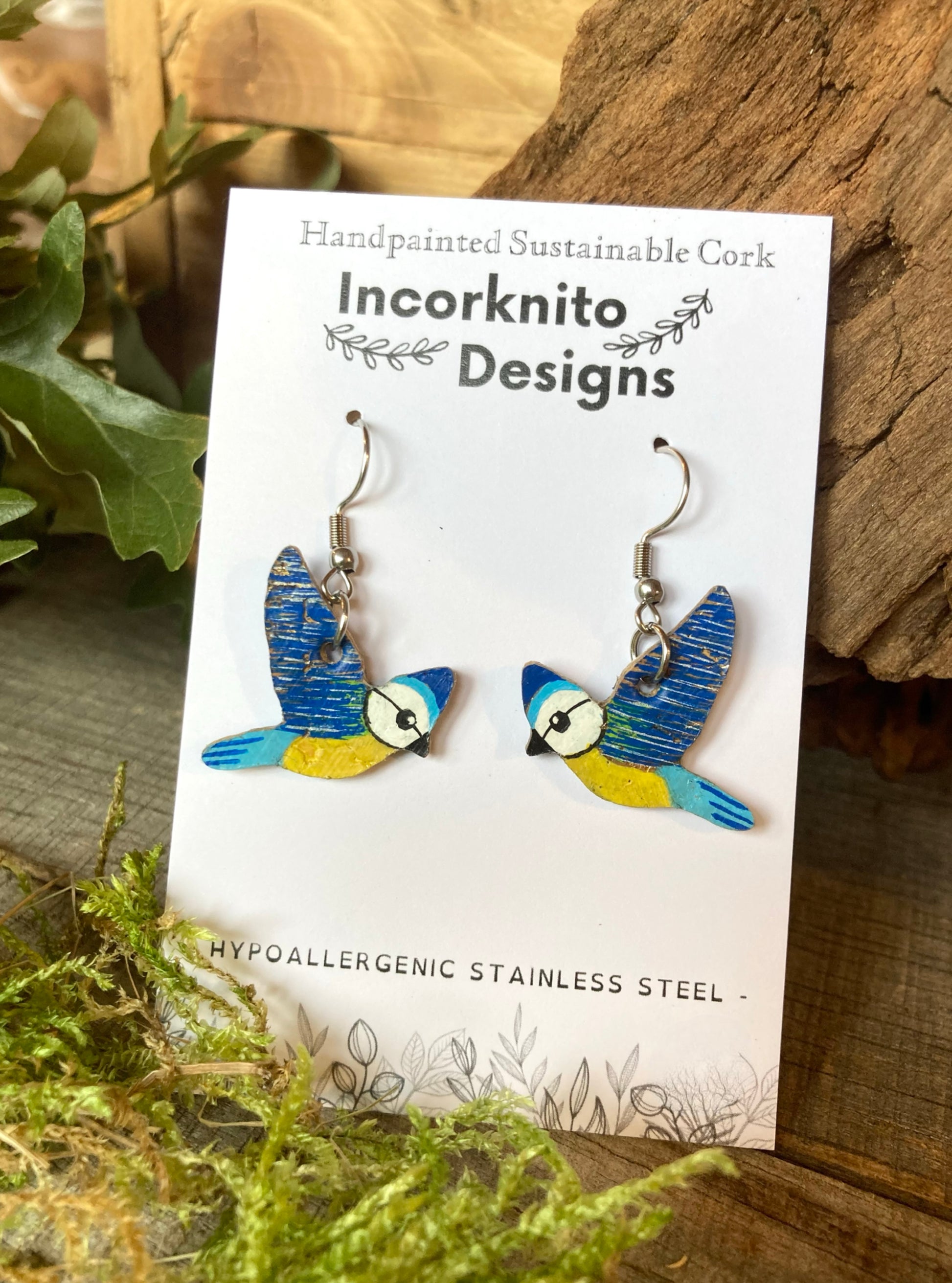 Bird-shaped earrings on a card with 'Incorknito Designs' branding, placed on a wooden surface with greenery.