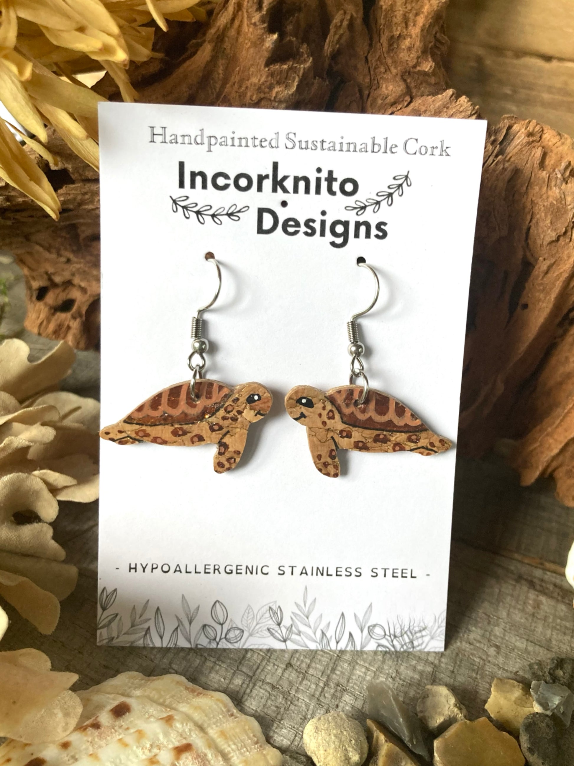 Handpainted turtle-shaped earrings on a card with 'Incorknito Designs' branding, surrounded by natural elements.