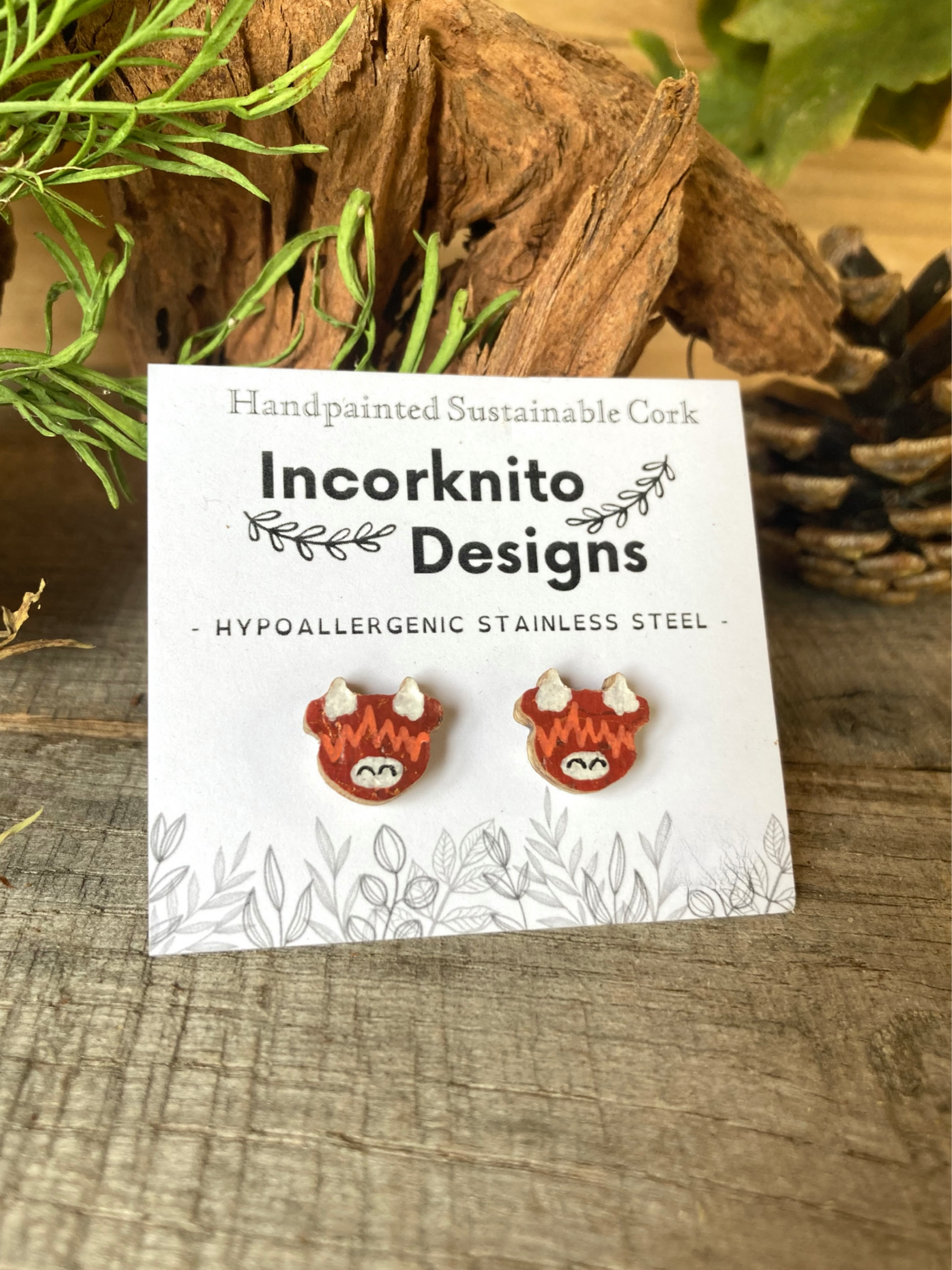 Pair of highland cow-shaped earrings on a card with 'Incorknito Designs' branding, set against a natural wood background.