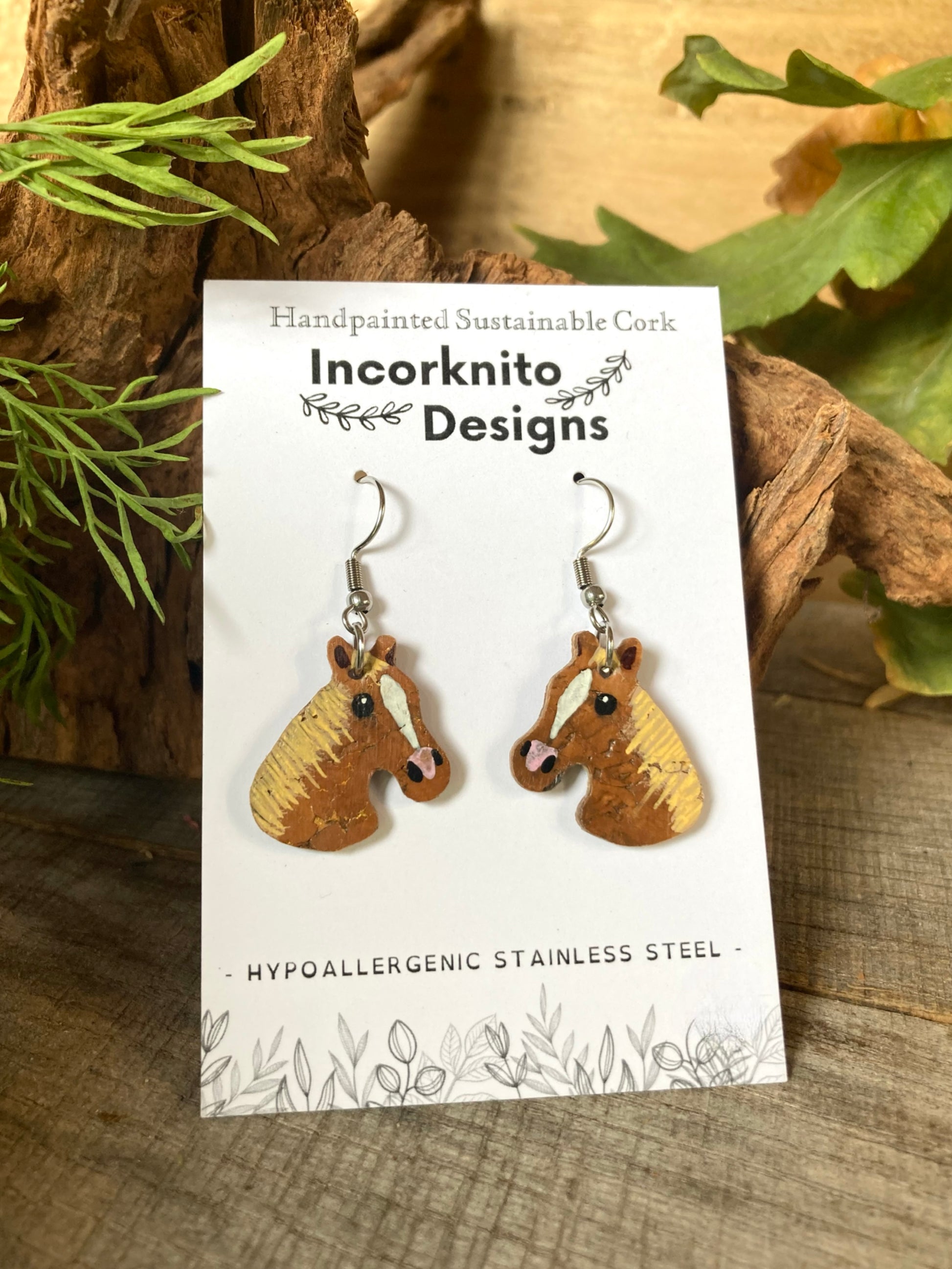 Horse-shaped earrings on a card with 'Incorknito Designs' branding, placed on a wooden surface with leaves.