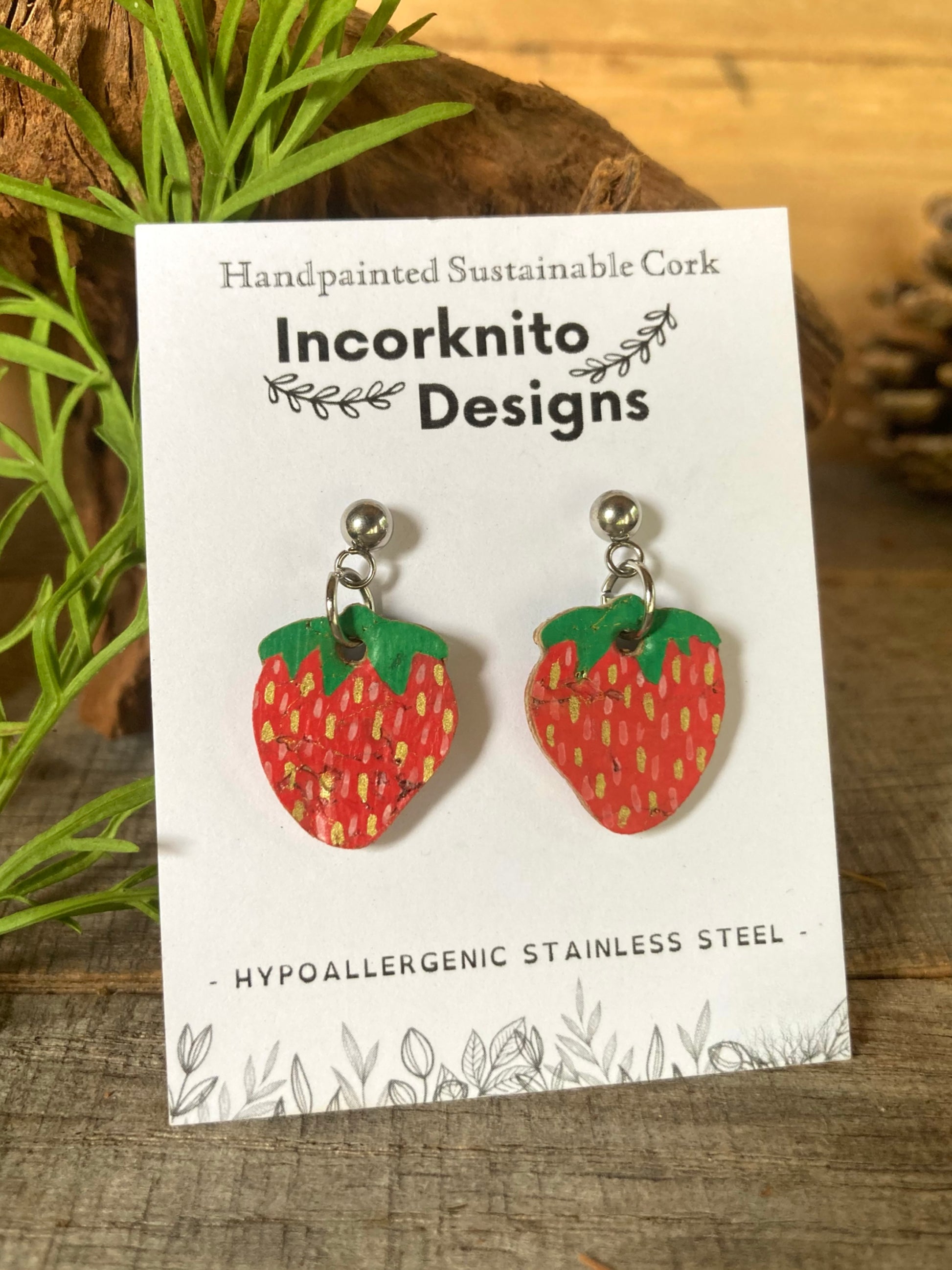 Strawberry-shaped earrings on a card with 'Incorknito Designs' branding, placed on a wooden surface.