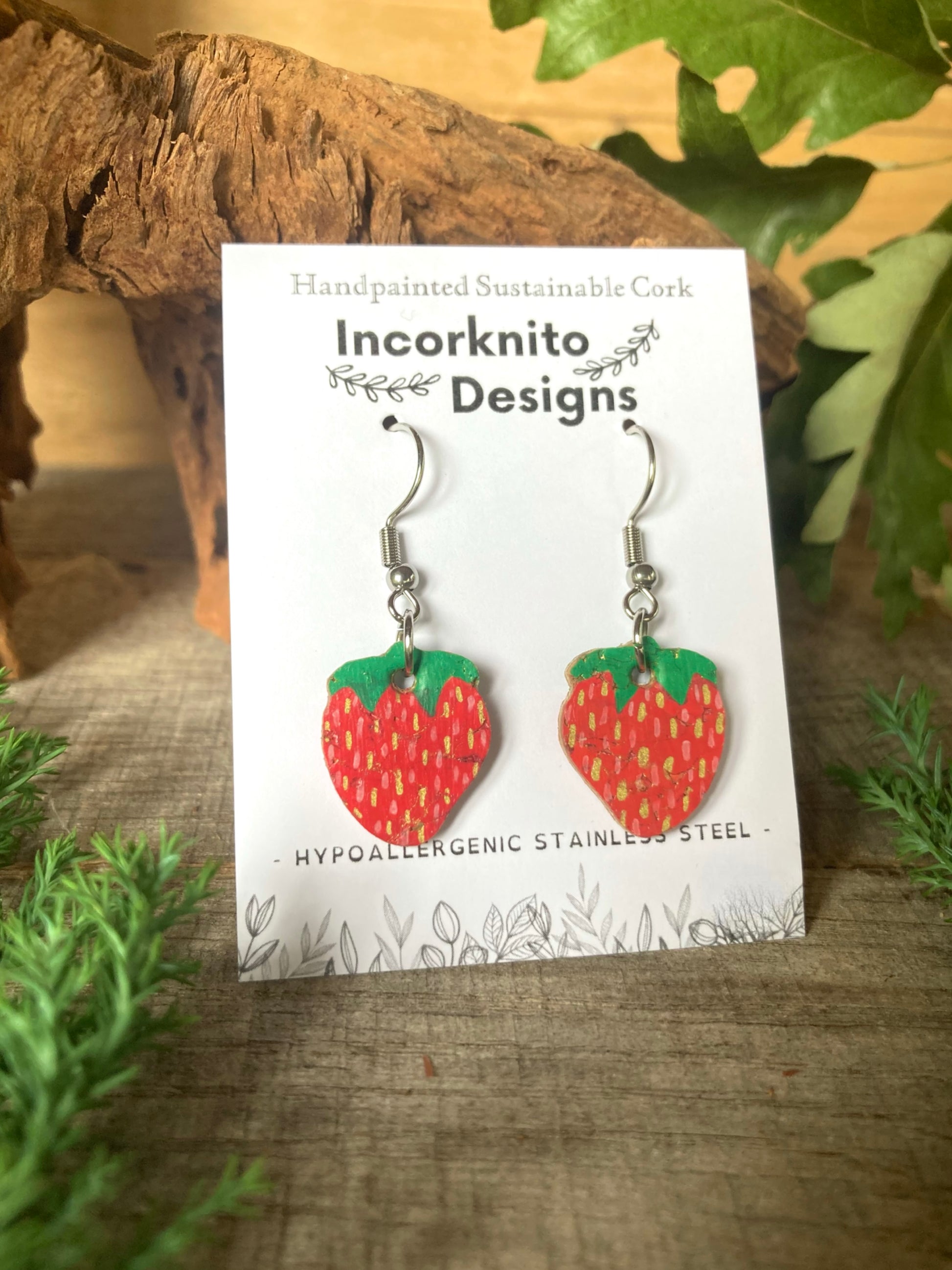 Strawberry-shaped earrings on a card with 'Incorknito Designs' branding, placed on a wooden surface with greenery.