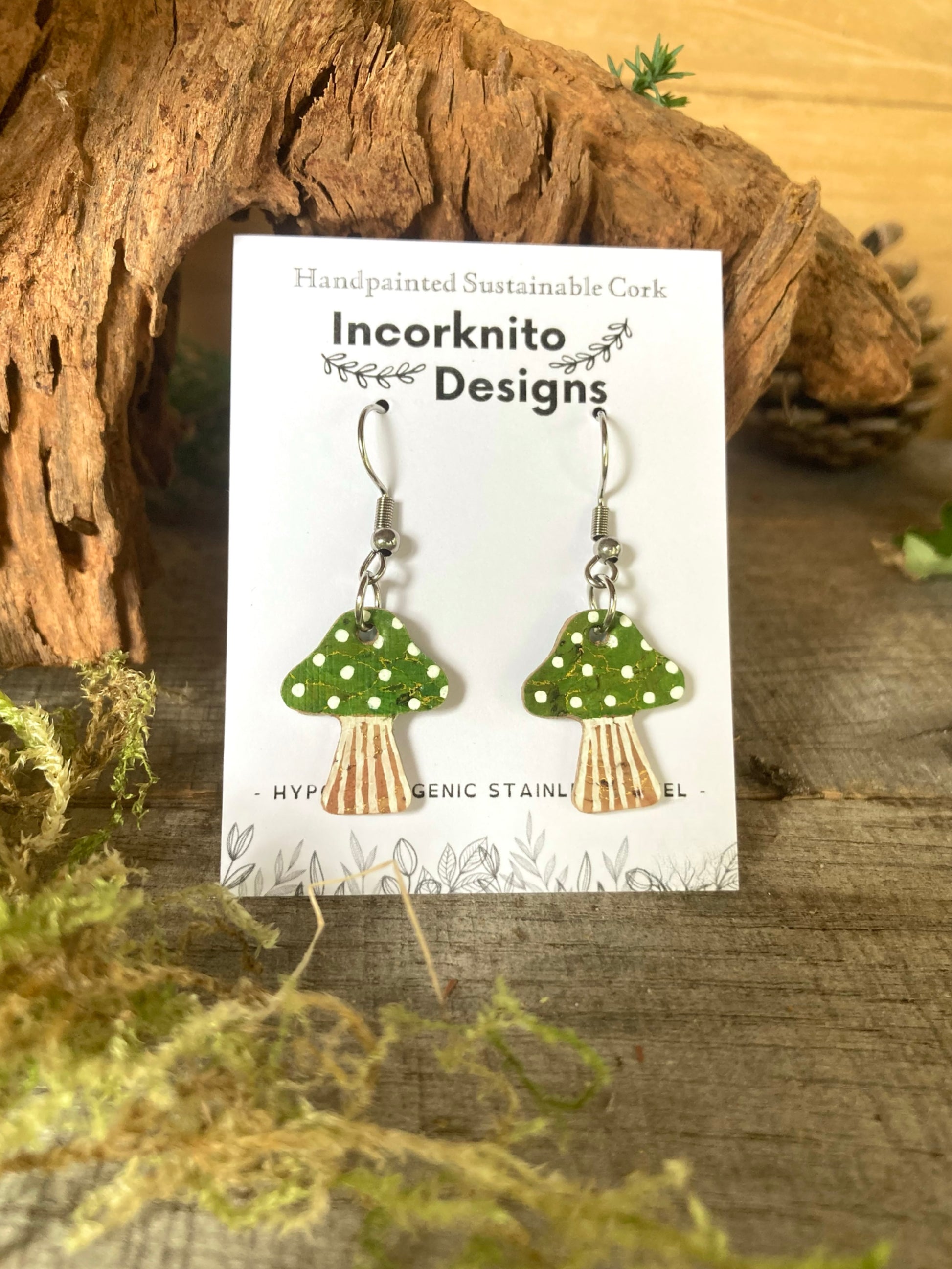 Green toadstool earrings on a backing card labelled 'Incorknito Designs' on a wooden surface
