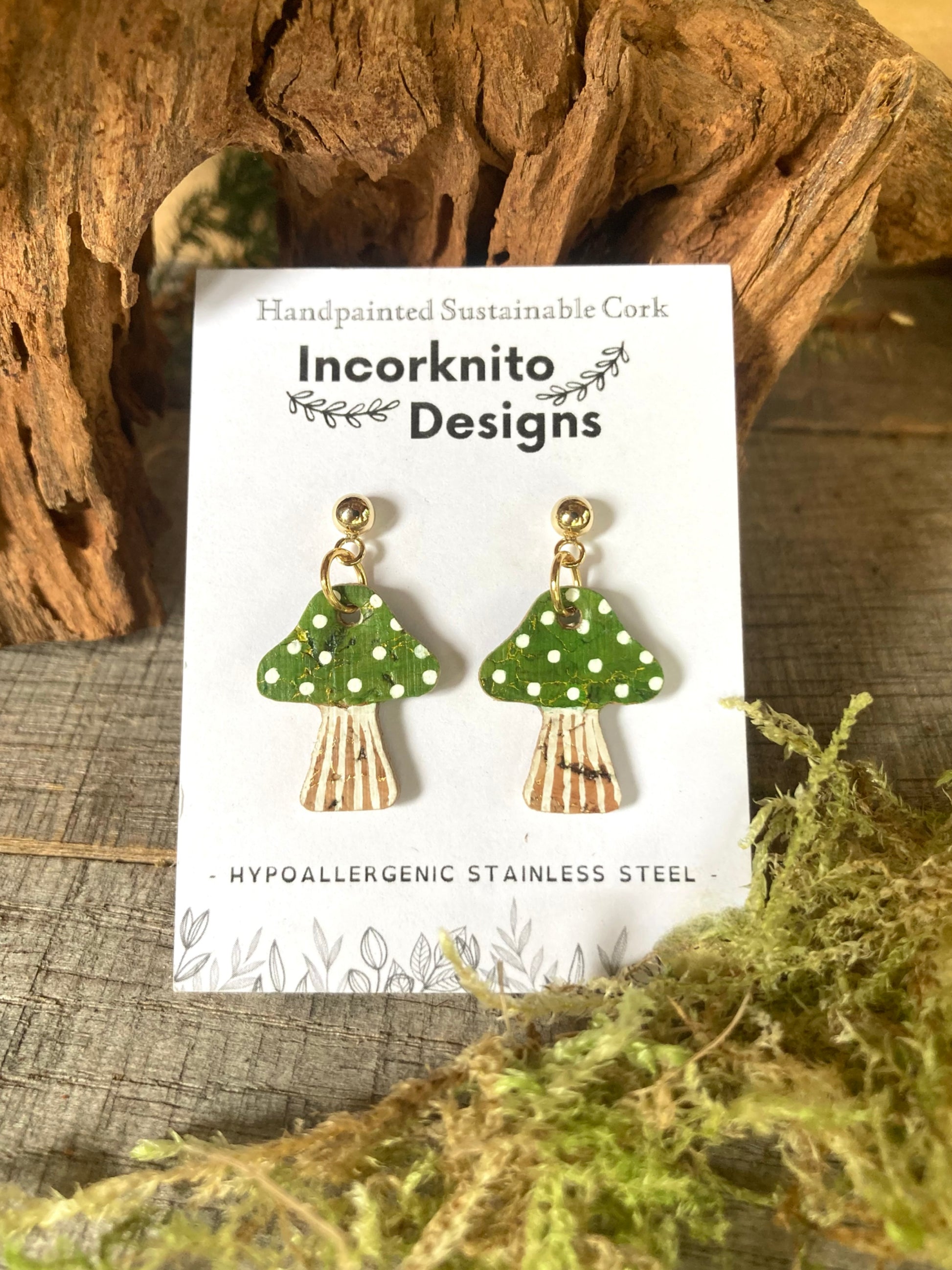Green mushroom-shaped earrings on a white card with 'Incorknito Designs' branding, placed on a wooden surface with moss.