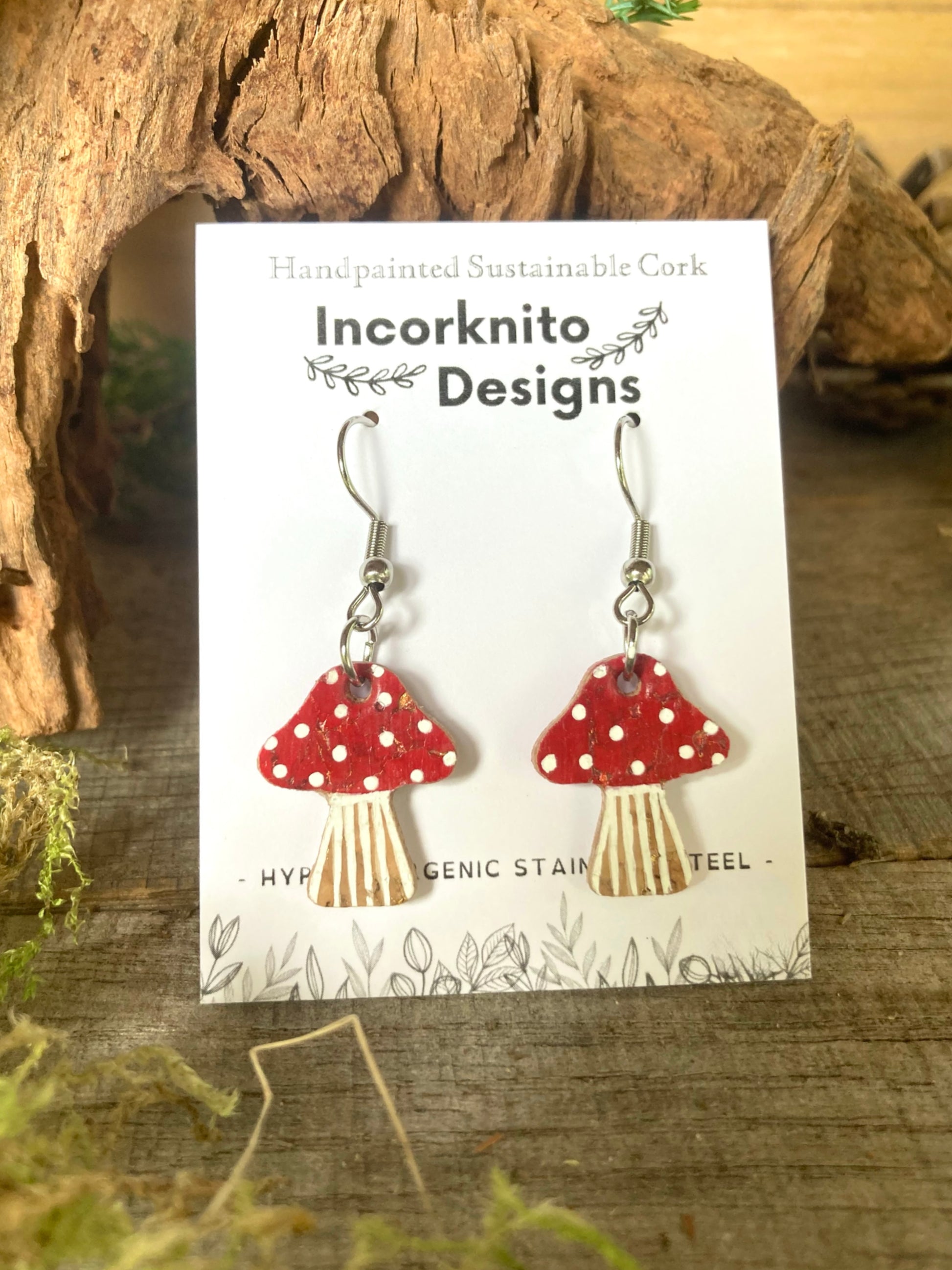 Red mushroom-shaped earrings with hooks on a 'Incorknito Designs' card with a natural wood background.