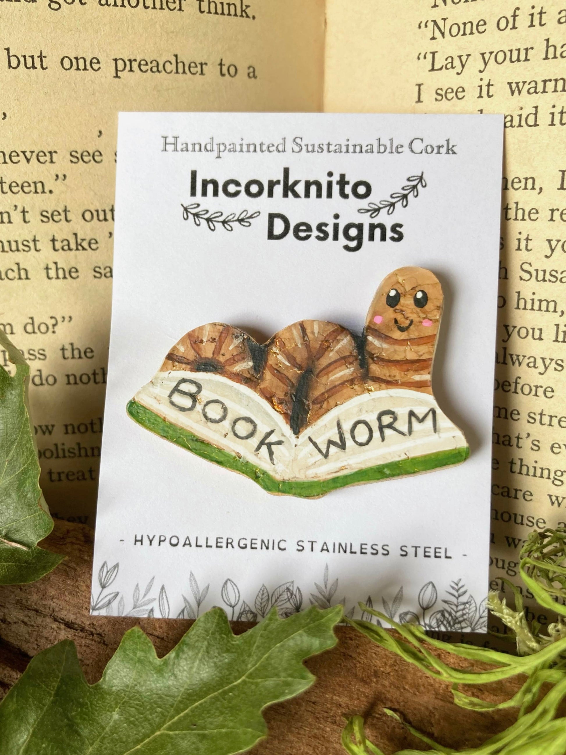 Pin with 'Book Worm' design on a card with 'Incorknito Designs' branding, set against a textured background.