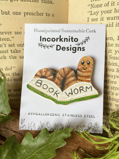 Pin with 'Book Worm' design on a card with 'Incorknito Designs' branding, set against a textured background.
