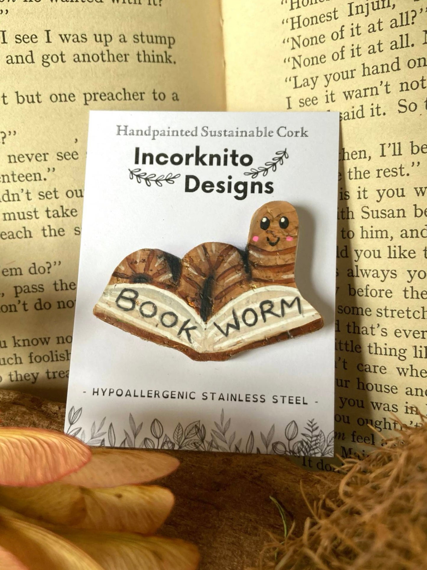 Bookworm-shaped pin with 'Incorknito Designs' packaging on a textured surface.