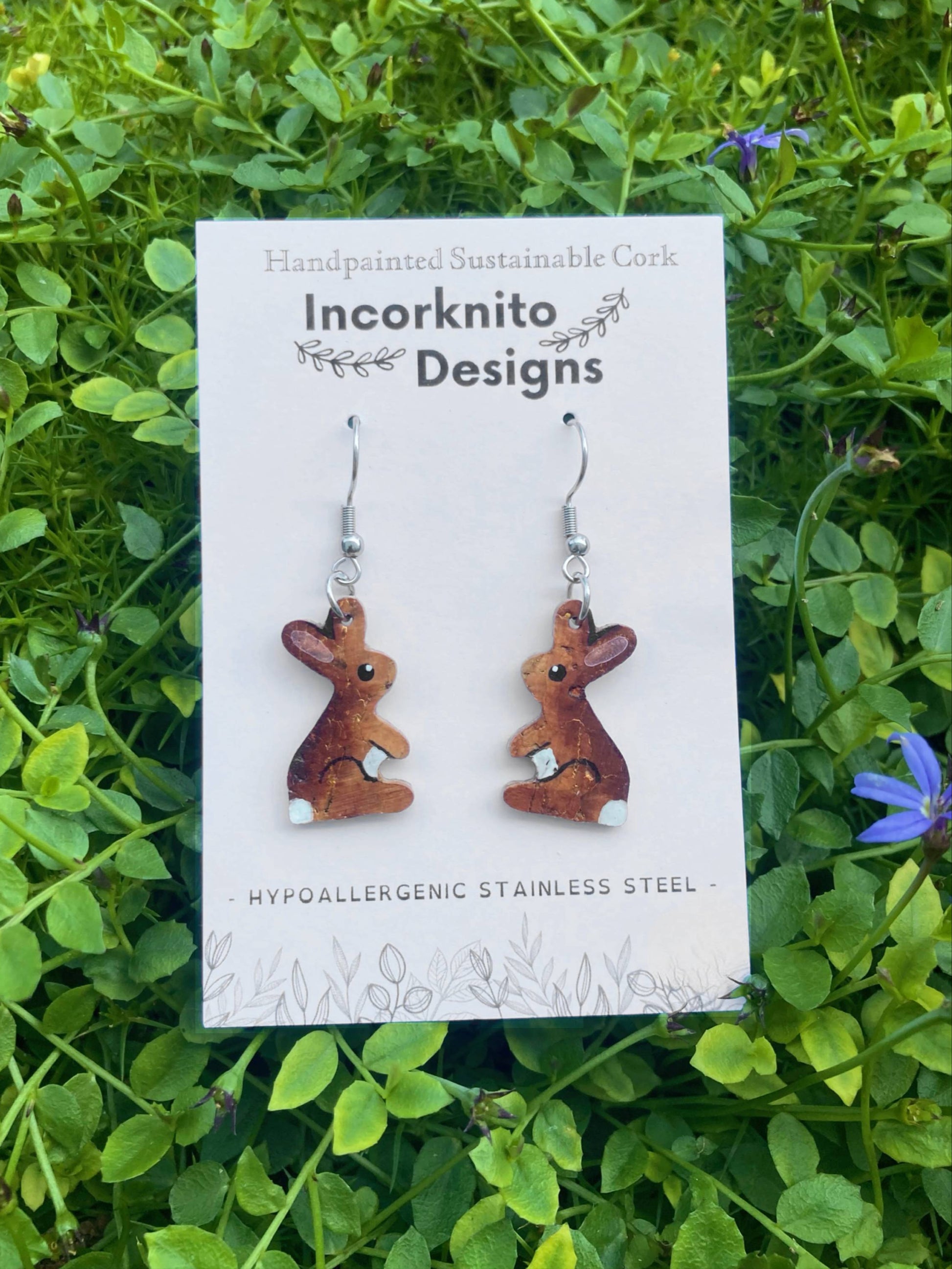 Pair of brown bunny-shaped earrings on a 'Incorknito Designs' card against a green grass background.