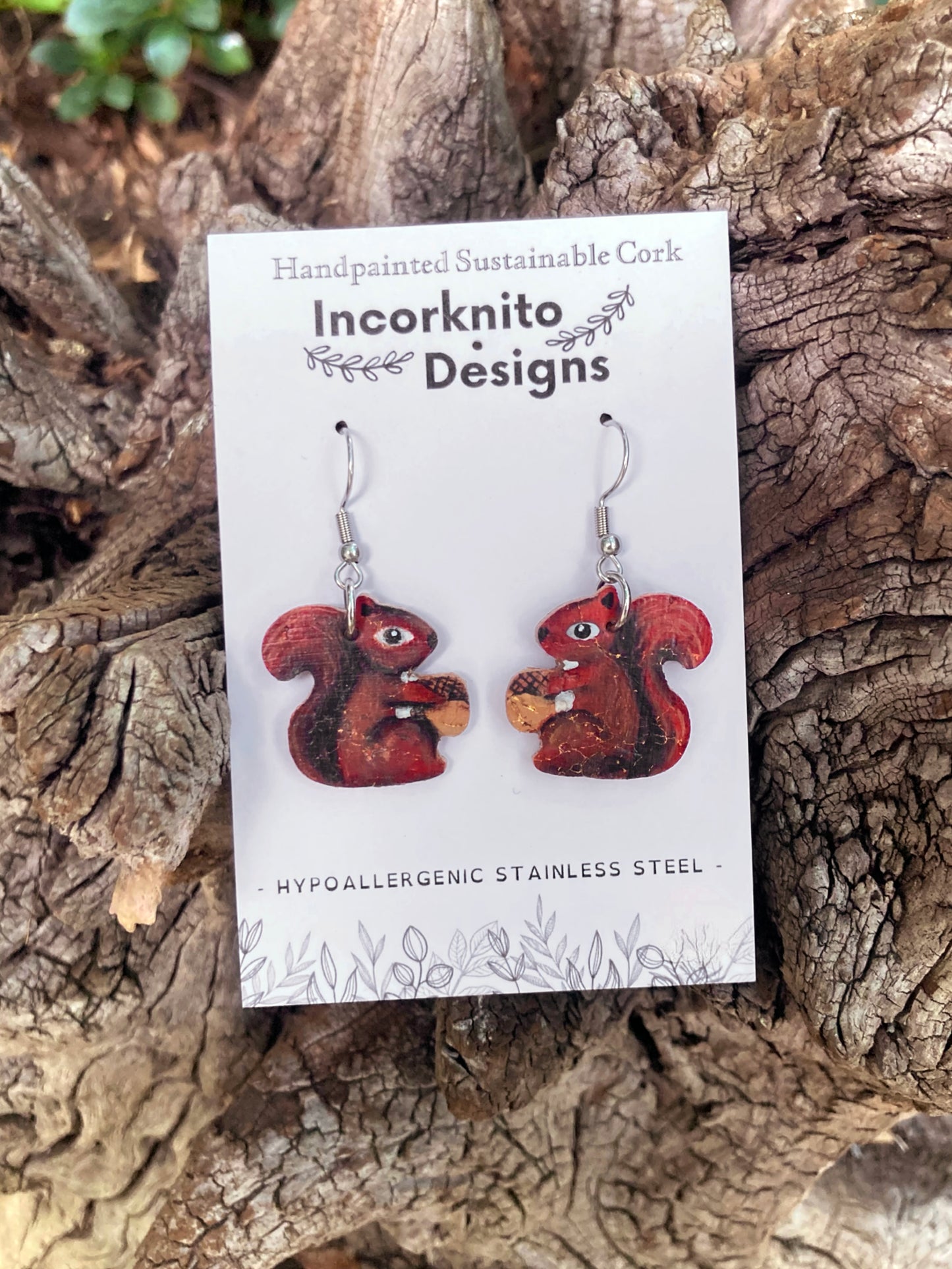 Red squirrel-shaped earrings on a card with 'Incorknito Designs' branding, placed on a wooden surface.