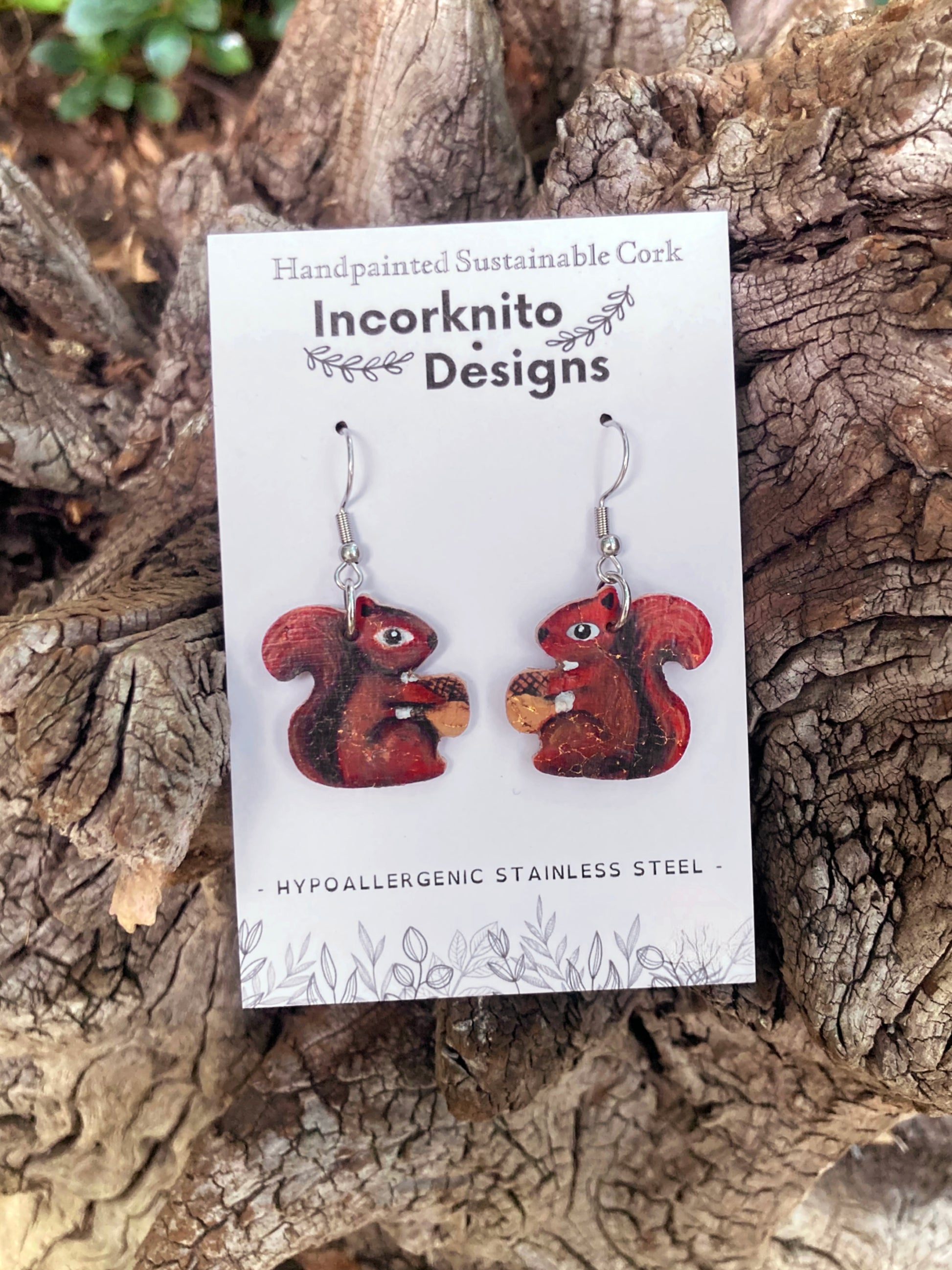 Red squirrel-shaped earrings on a card with 'Incorknito Designs' branding, placed on a wooden surface.