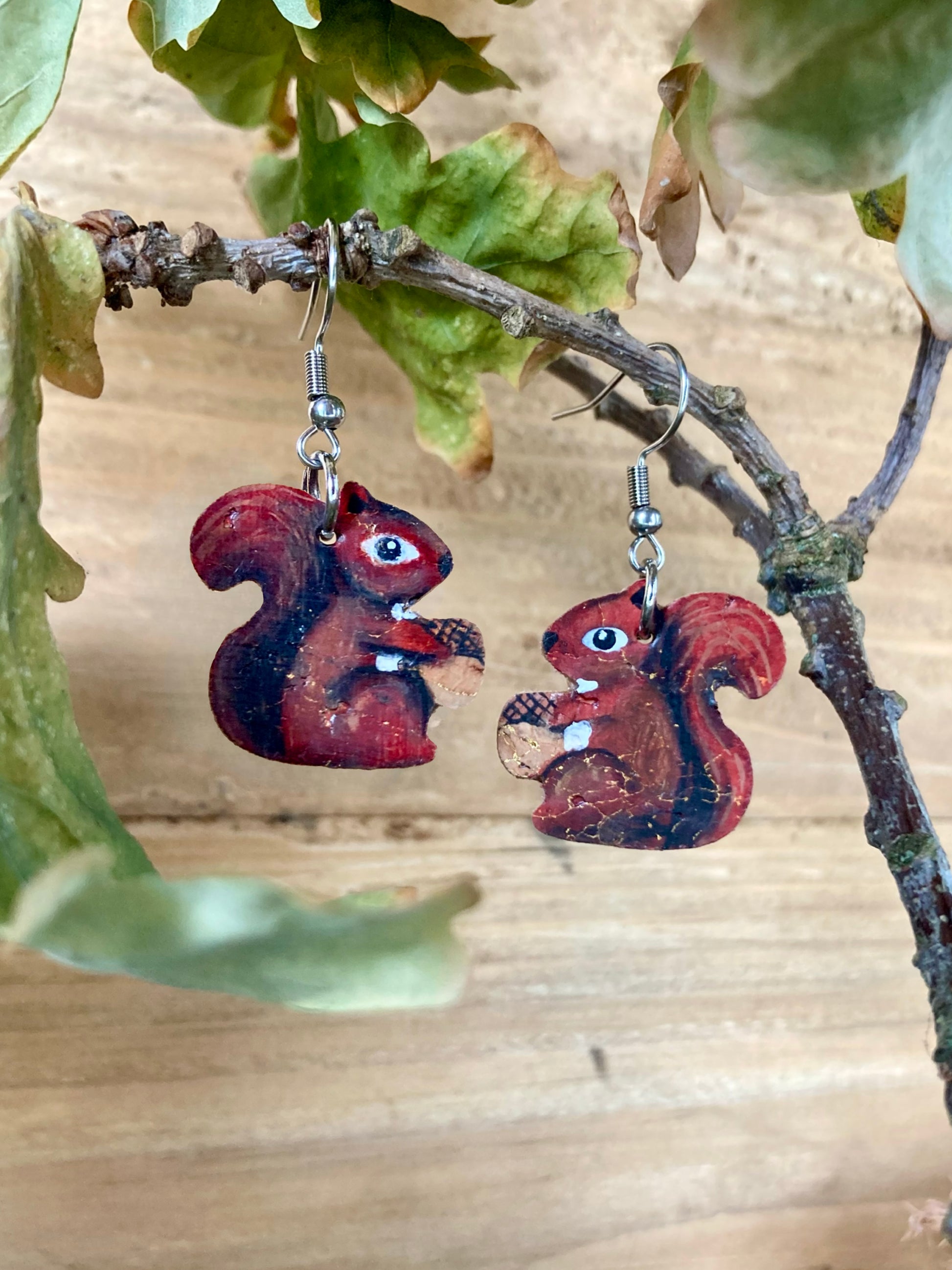 Red squirrel holding an acorn shaped earrings hanging from a branch with leaves.