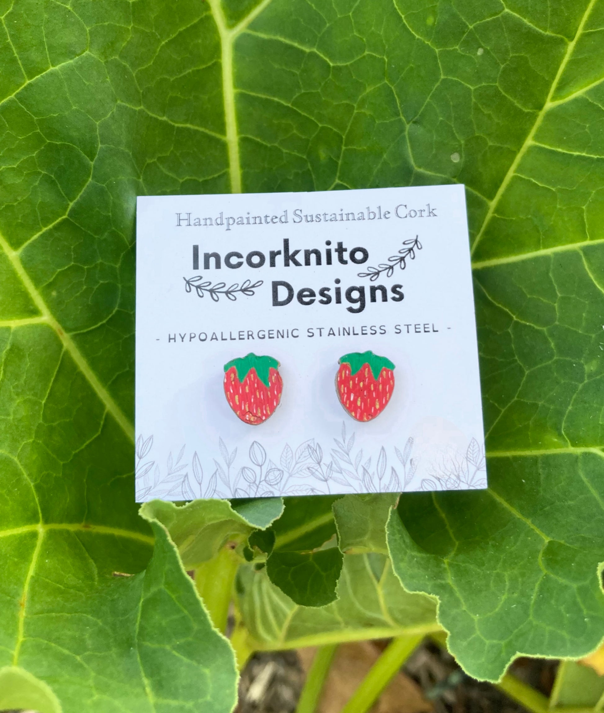 Strawberry earrings on a white card with 'Incorknito Designs' branding, set against a green leafy background.