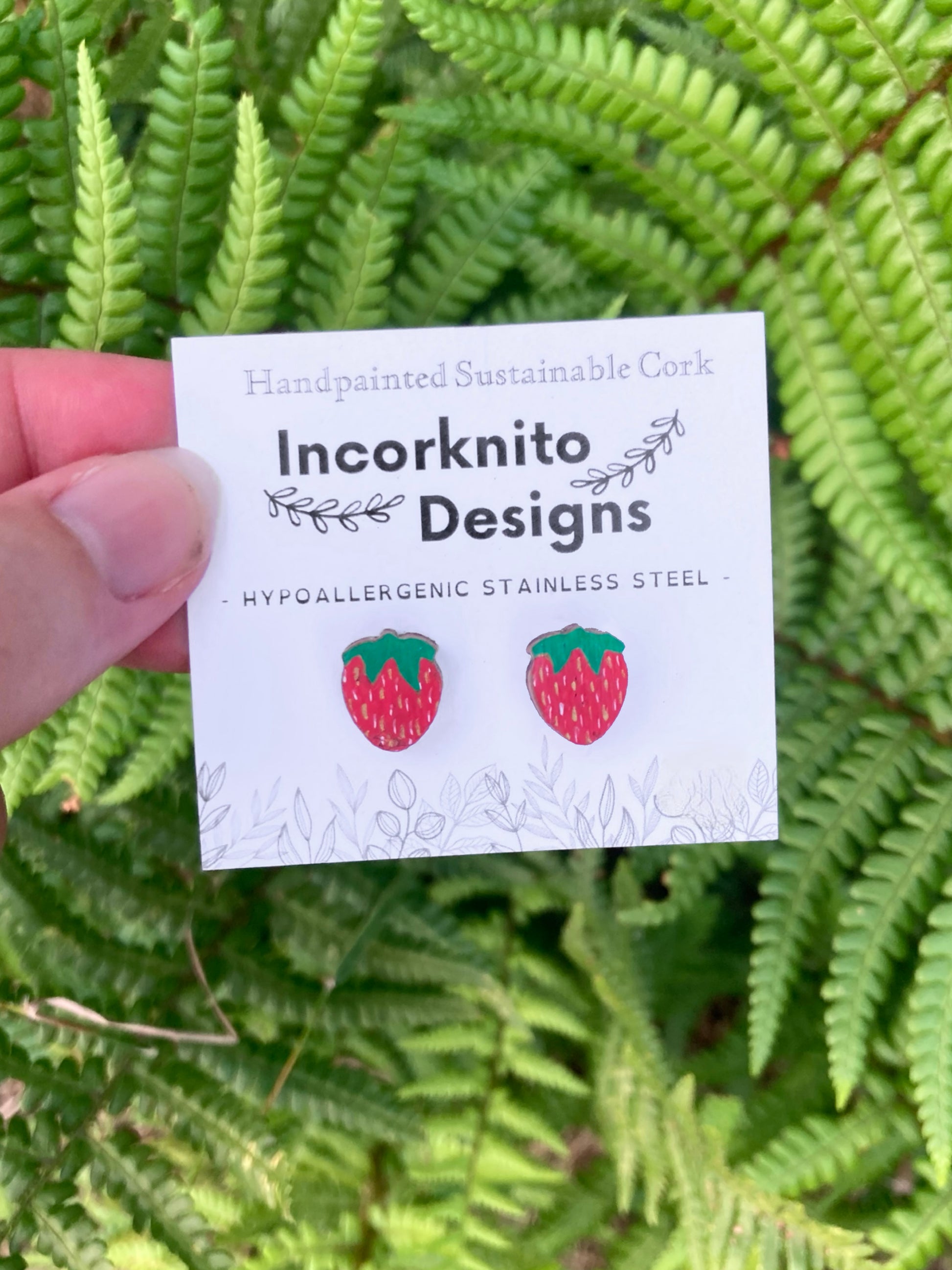 Card with strawberry earrings design held over green ferns