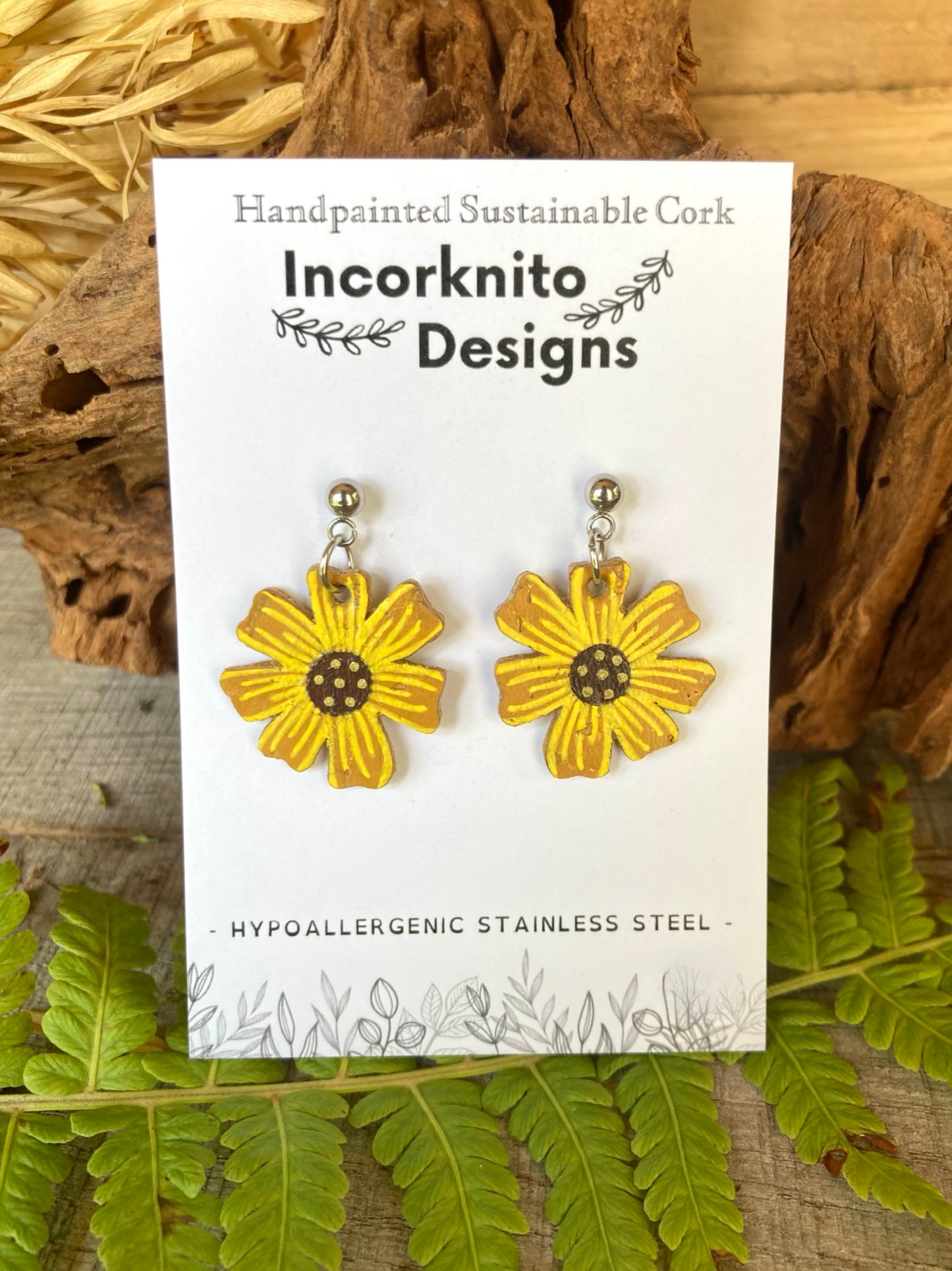 Gold flower-shaped earrings on a white card with 'Incorknito Designs' branding, surrounded by natural elements.