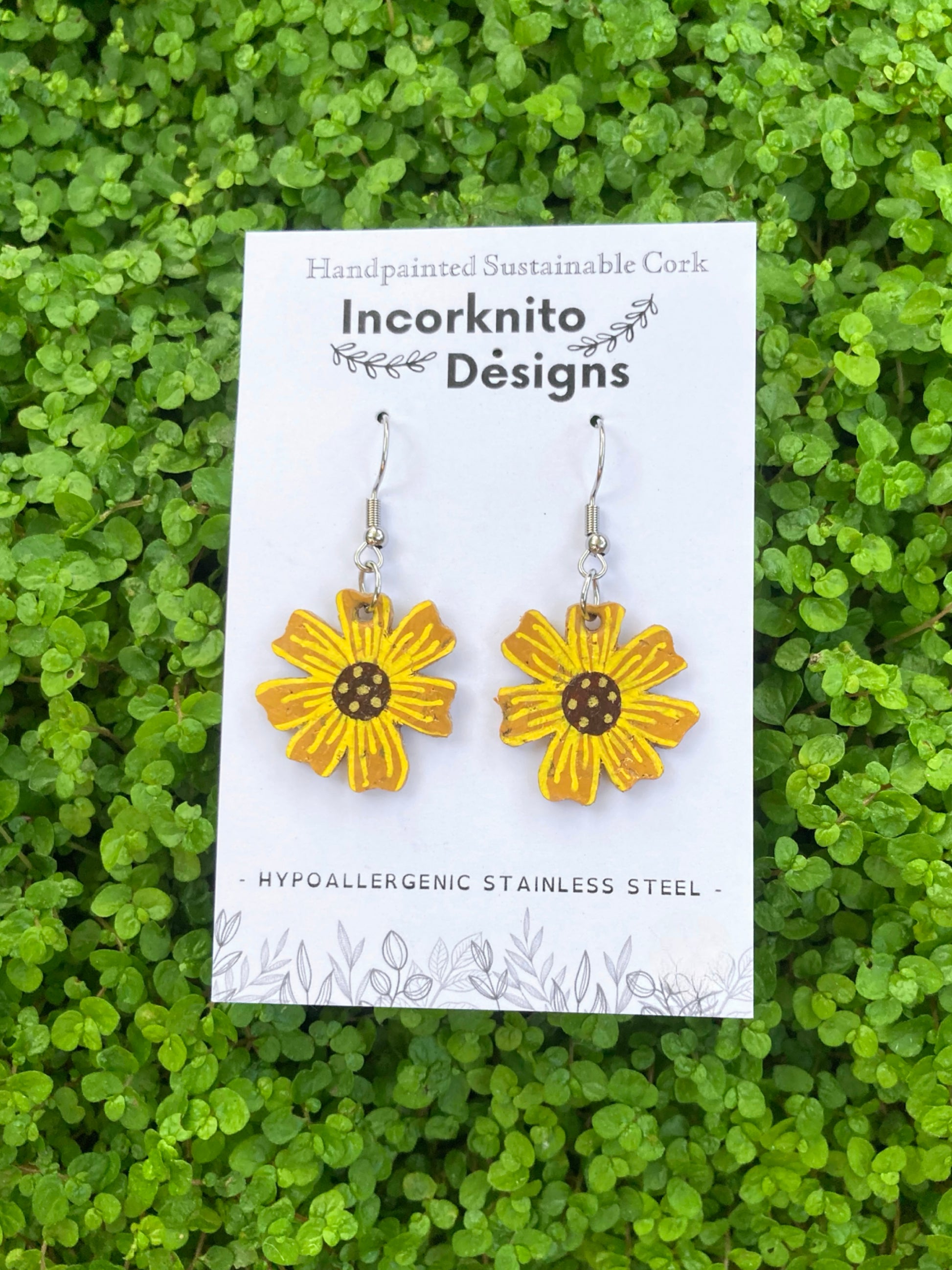 Yellow flower-shaped earrings on a white card with 'Incorknito Designs' branding, set against a green leafy background.