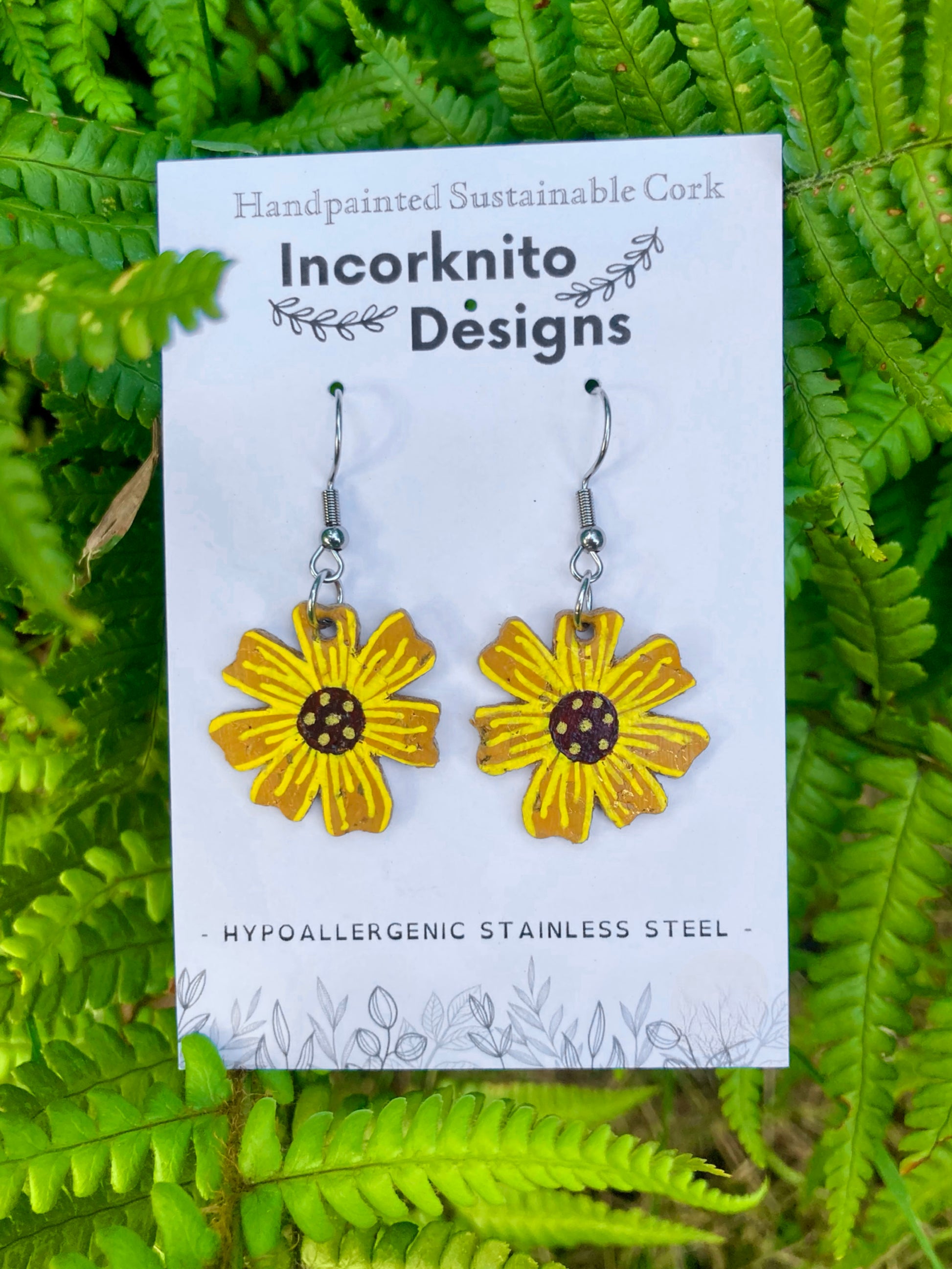 Yellow flower-shaped earrings on a white card with 'Incorknito Designs' branding, set against a green leafy background.
