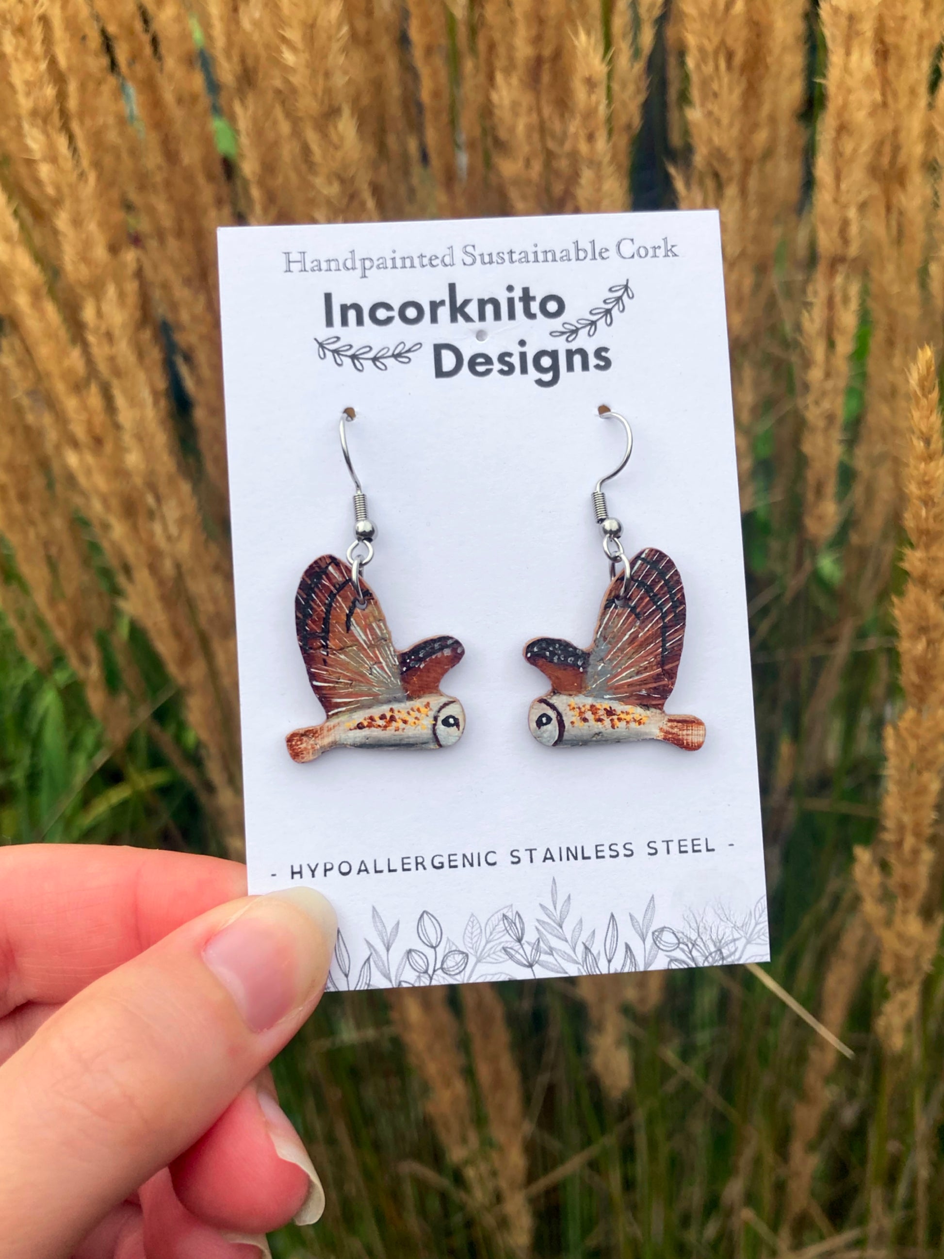 Owl earrings on hooks on 'Incorknito Designs' backing card being held in front of tall grass