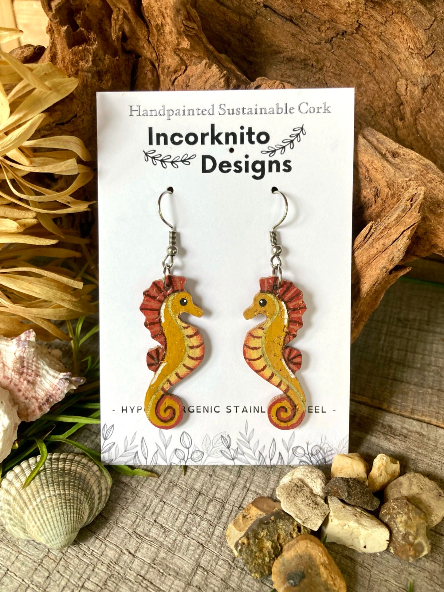 Handpainted sustainable cork seahorse earrings on a white card with 'Incorknito Designs' branding, surrounded by natural elements.