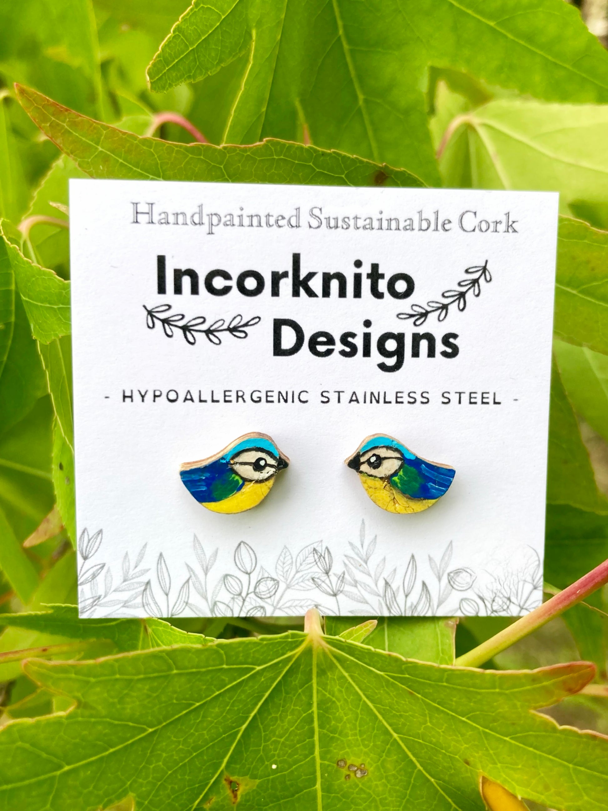 Blue-tit shaped earrings on a card with 'Incorknito Designs' branding, placed on green leaves.