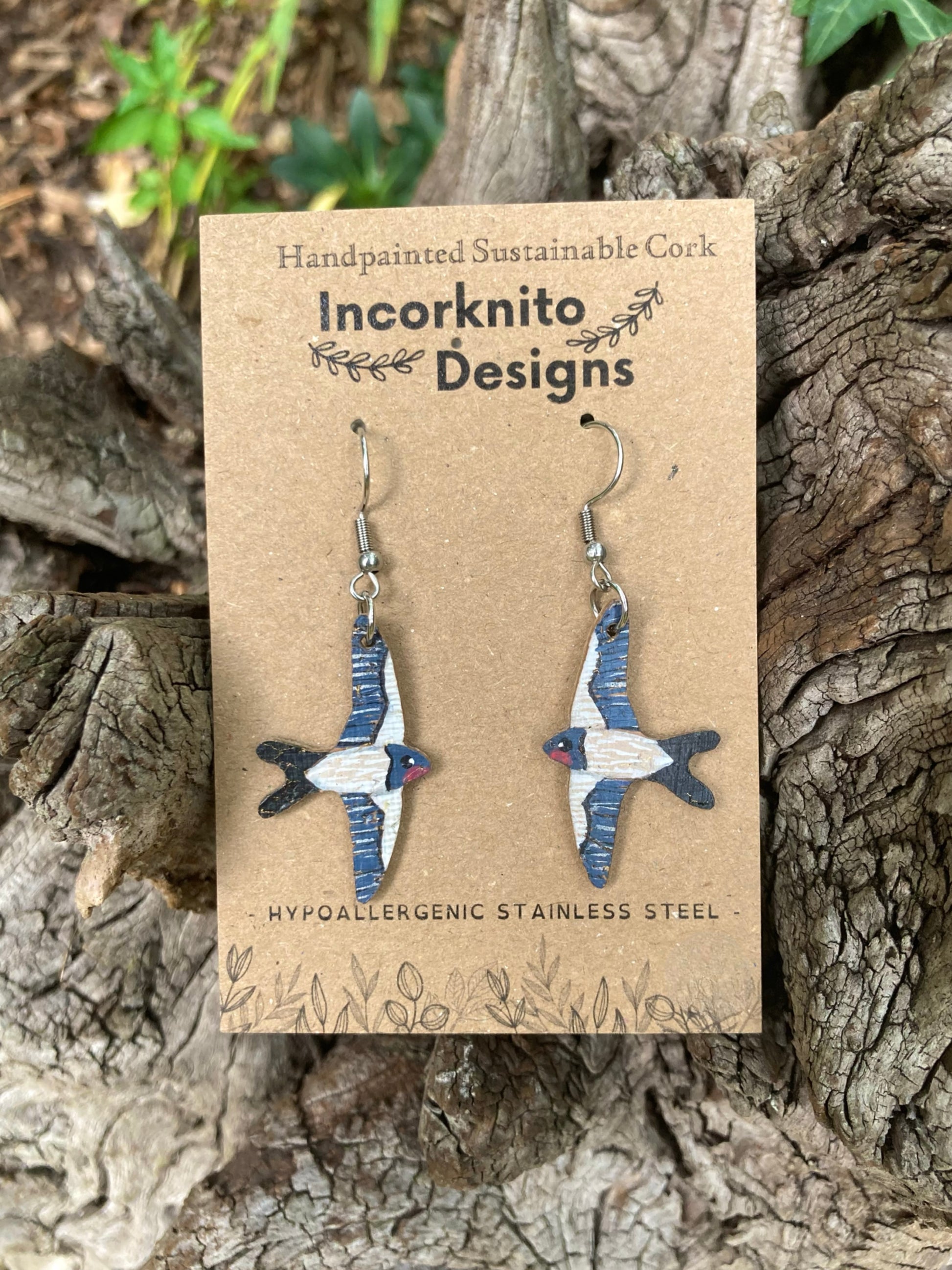 Flying swallow bird-shaped earrings on a card with 'Incorknito Designs' branding, placed on a wooden surface.